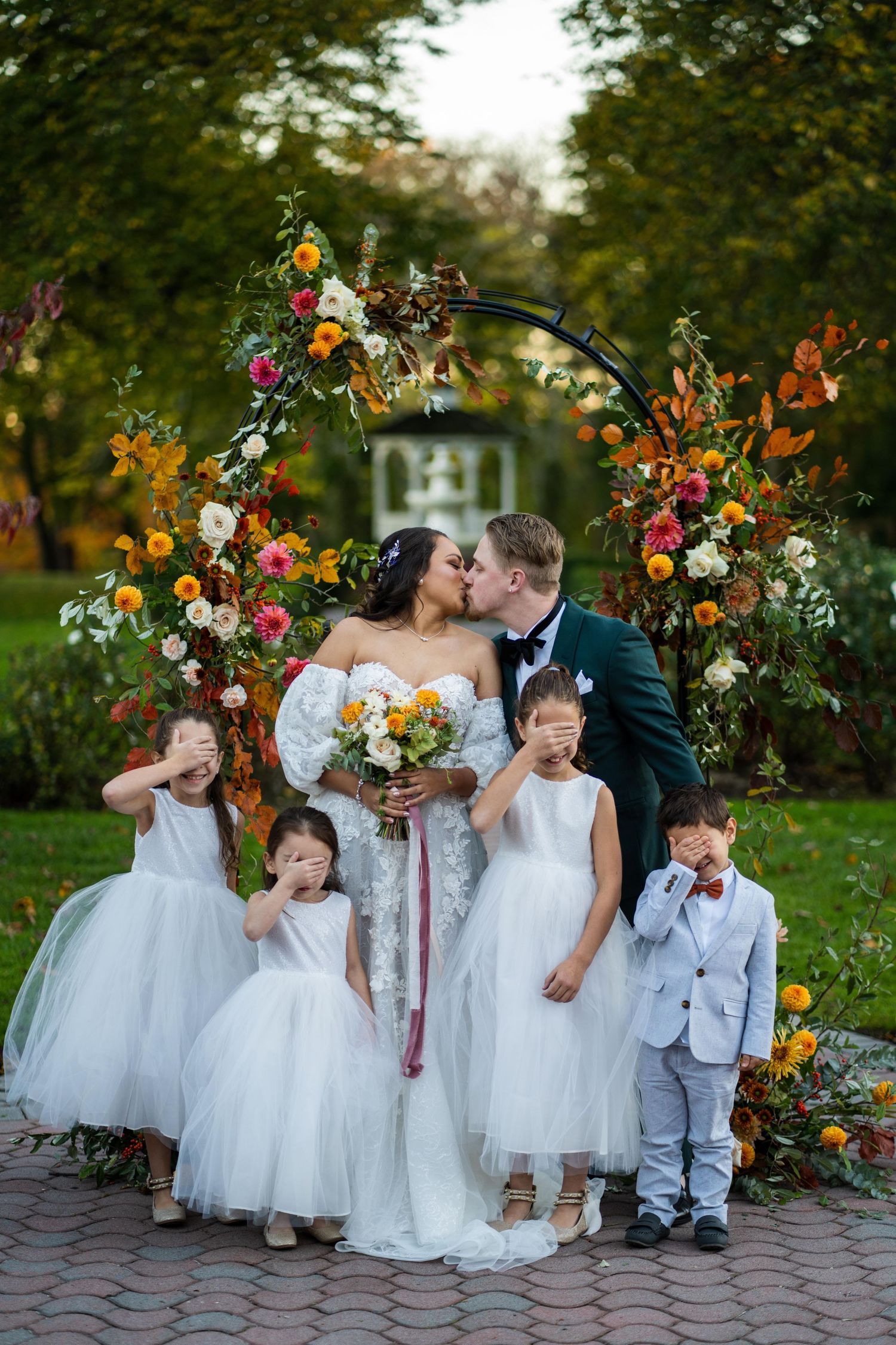 Wedding party with flower girls in white dresses posing by colorful floral arrangements.
