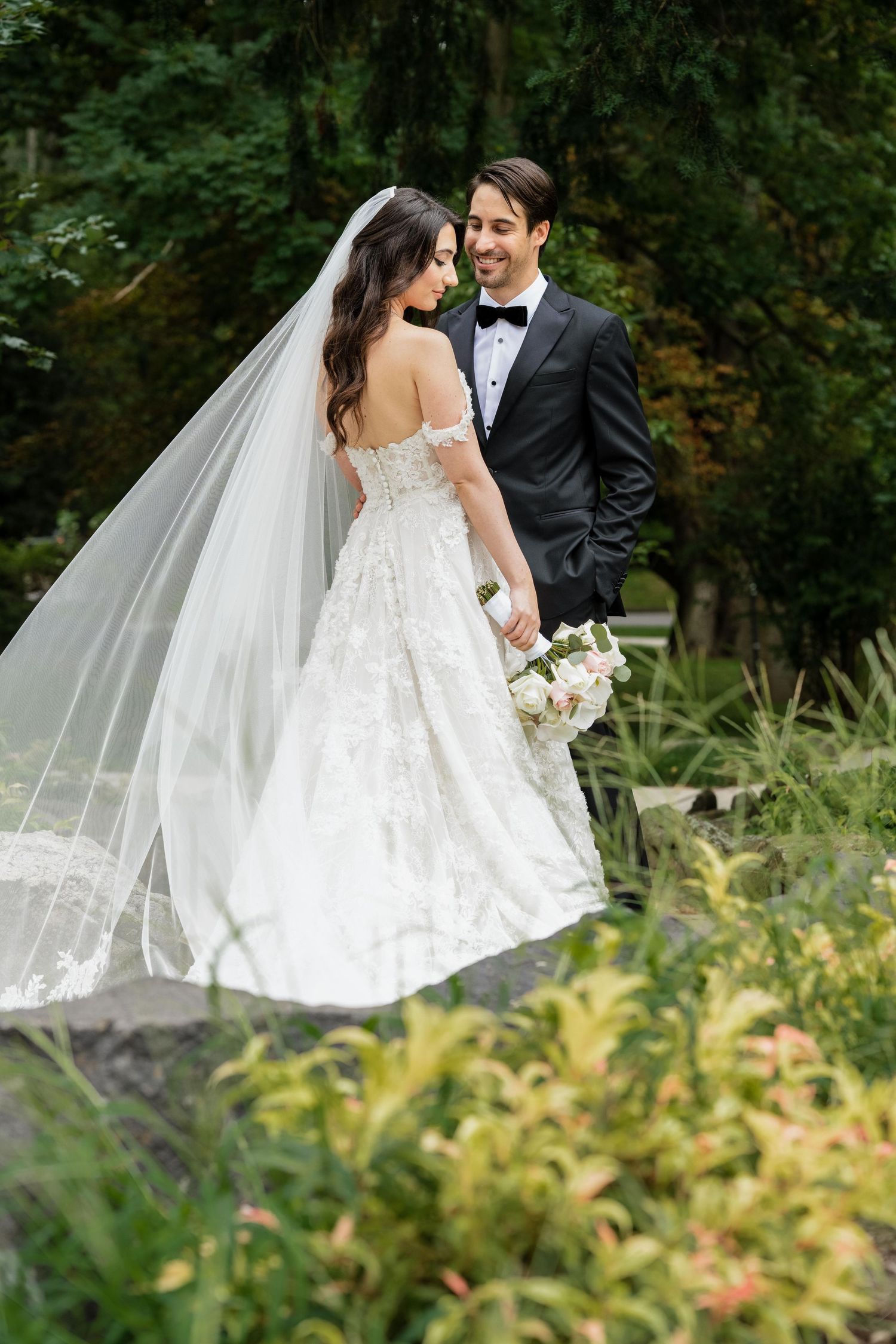 Bride in lace off-shoulder gown with veil holds bouquet beside groom in black tuxedo in lush garden setting.