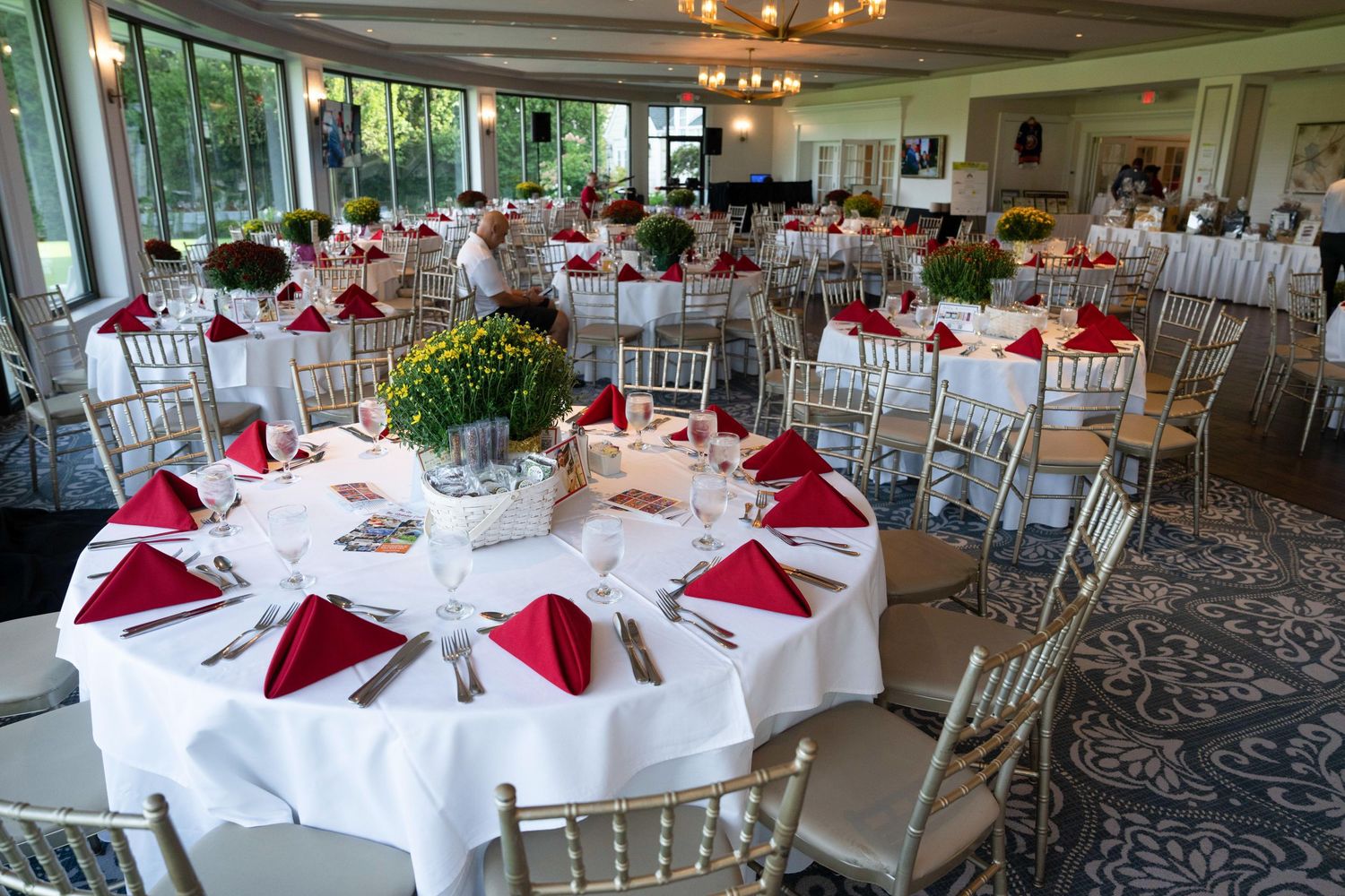 Elegant banquet hall with round tables, white linens, red napkins, and gold chiavari chairs for a formal event.