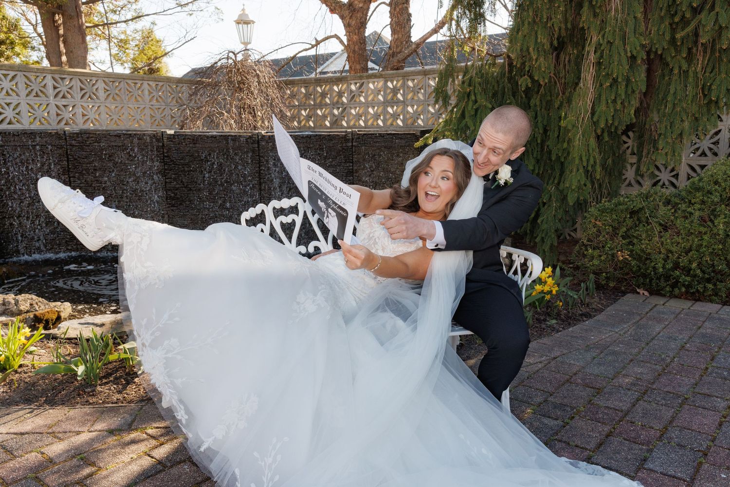 Groom in black tuxedo joyfully lifts smiling bride in white wedding gown outdoors in a garden setting.
