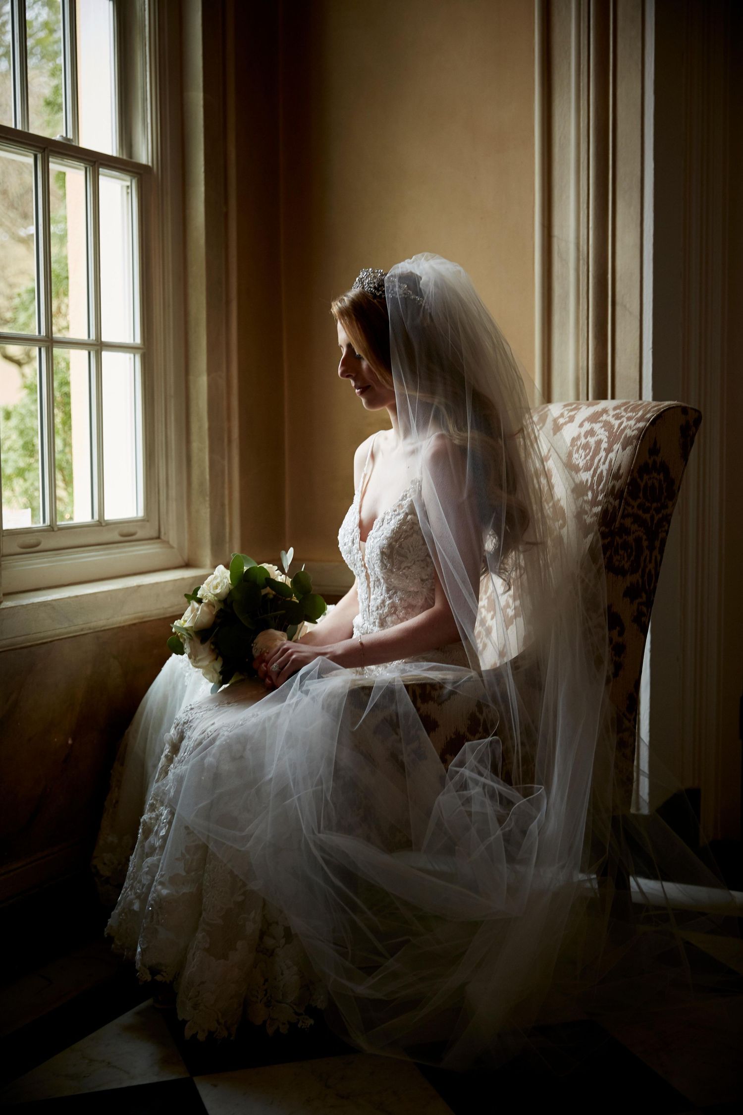 Bride in white wedding dress sits by window in dramatic lighting.