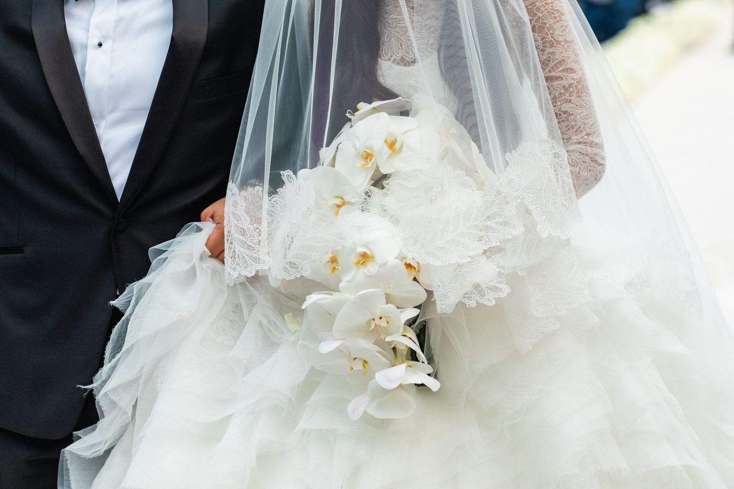 Bride in white ruffled gown holding white orchid bouquet beside groom in black tuxedo on wedding day.