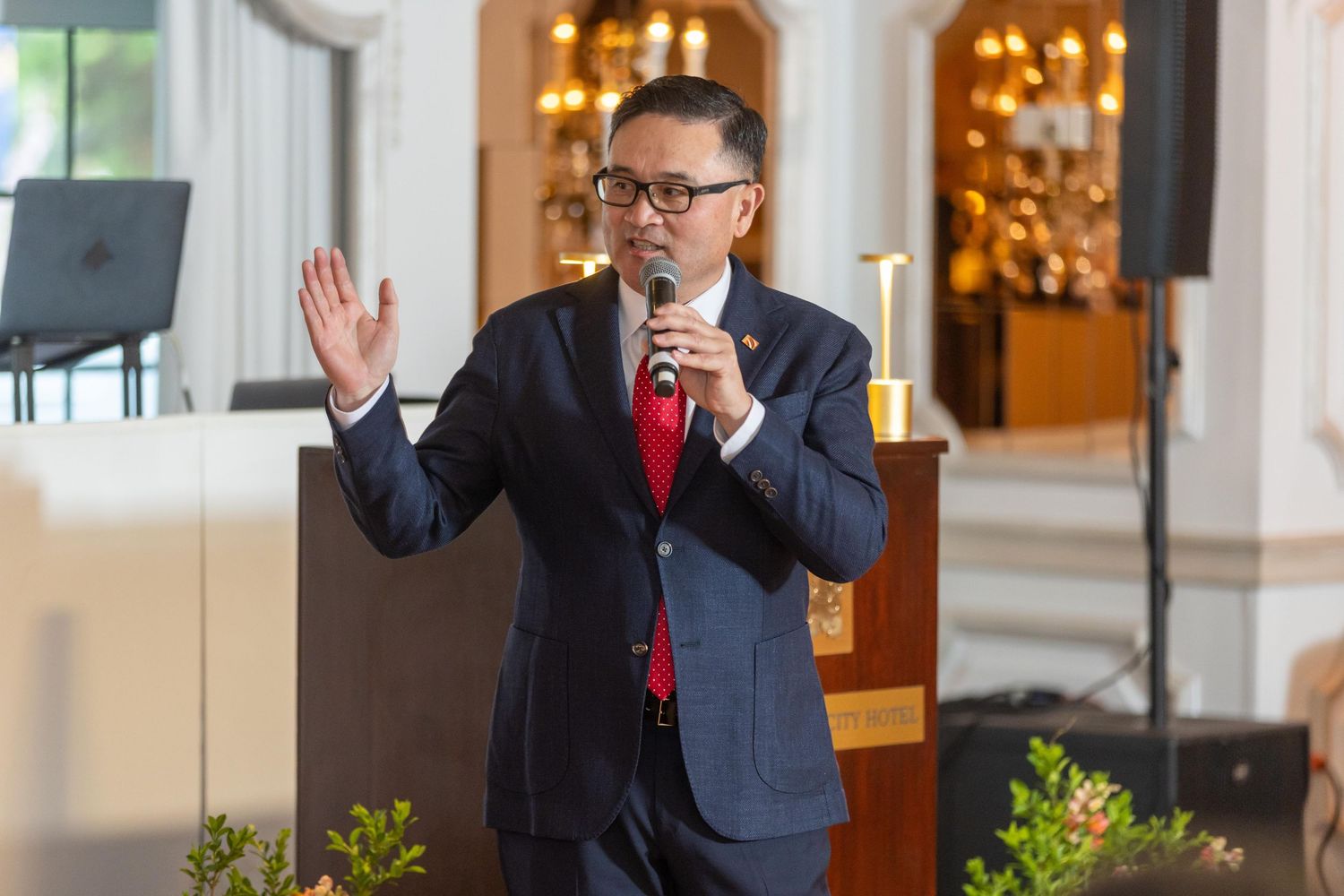 Man in navy suit speaking at podium with microphone, gesturing with raised hand at formal indoor event.