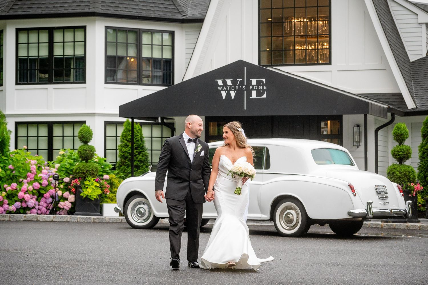 Elegant wedding couple poses by vintage white cars outside a grand estate building.