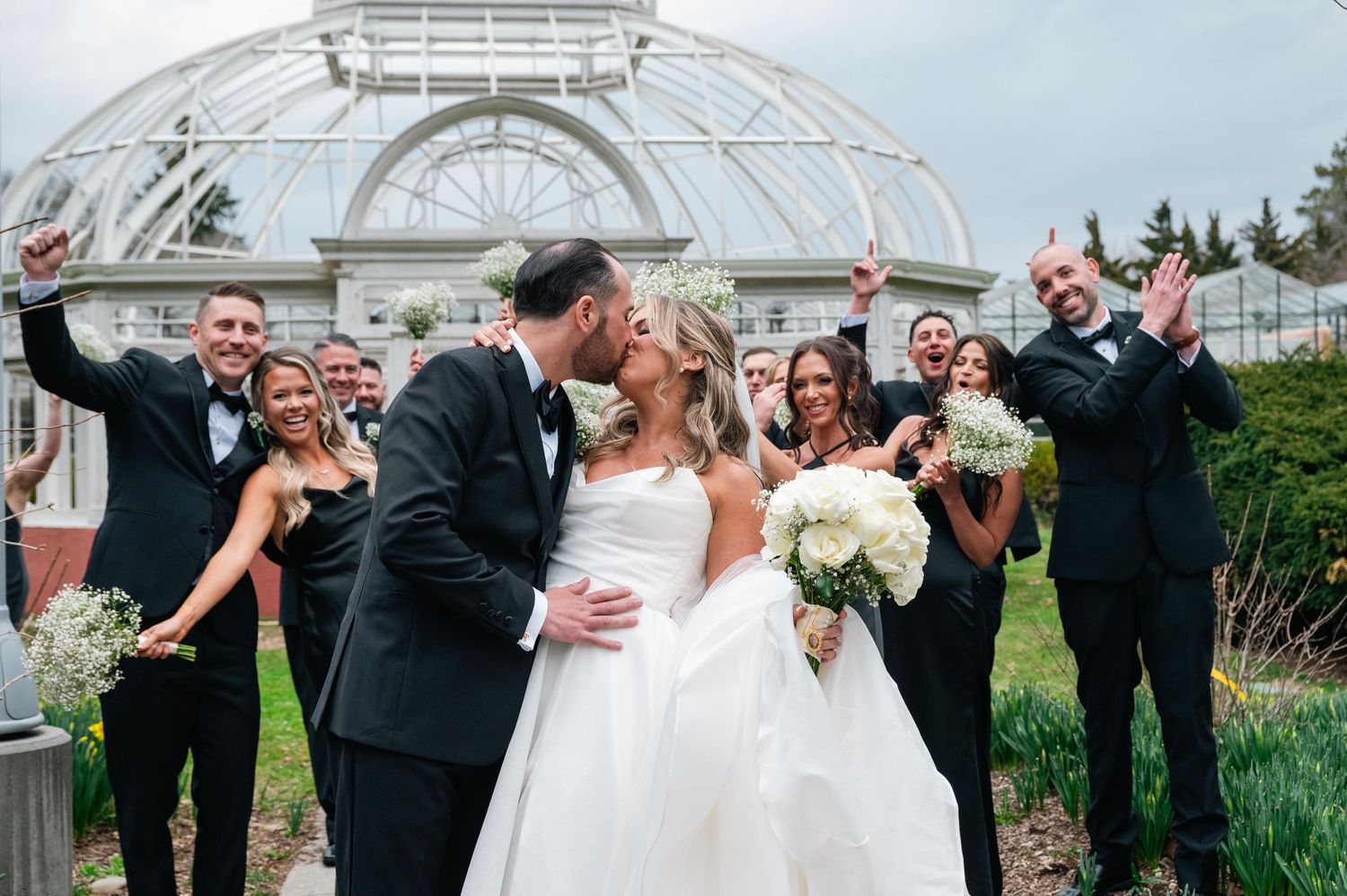 Wedding couple shares romantic kiss under garden gazebo surrounded by celebrating wedding party.