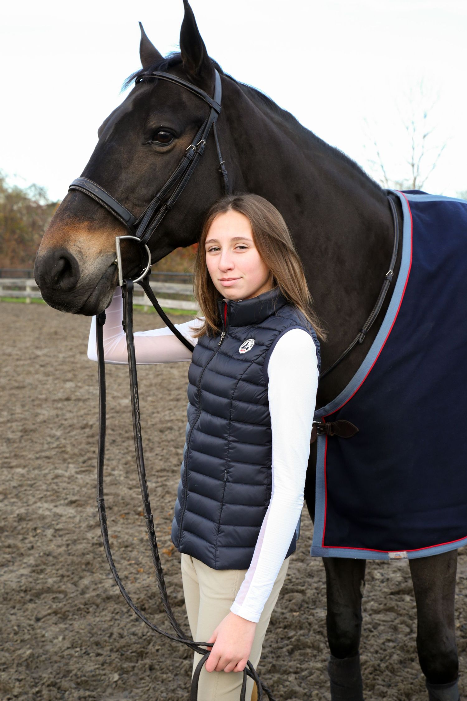 A rider in a navy puffer vest and white long sleeve shirt stands next to a dark horse wearing a blanket.