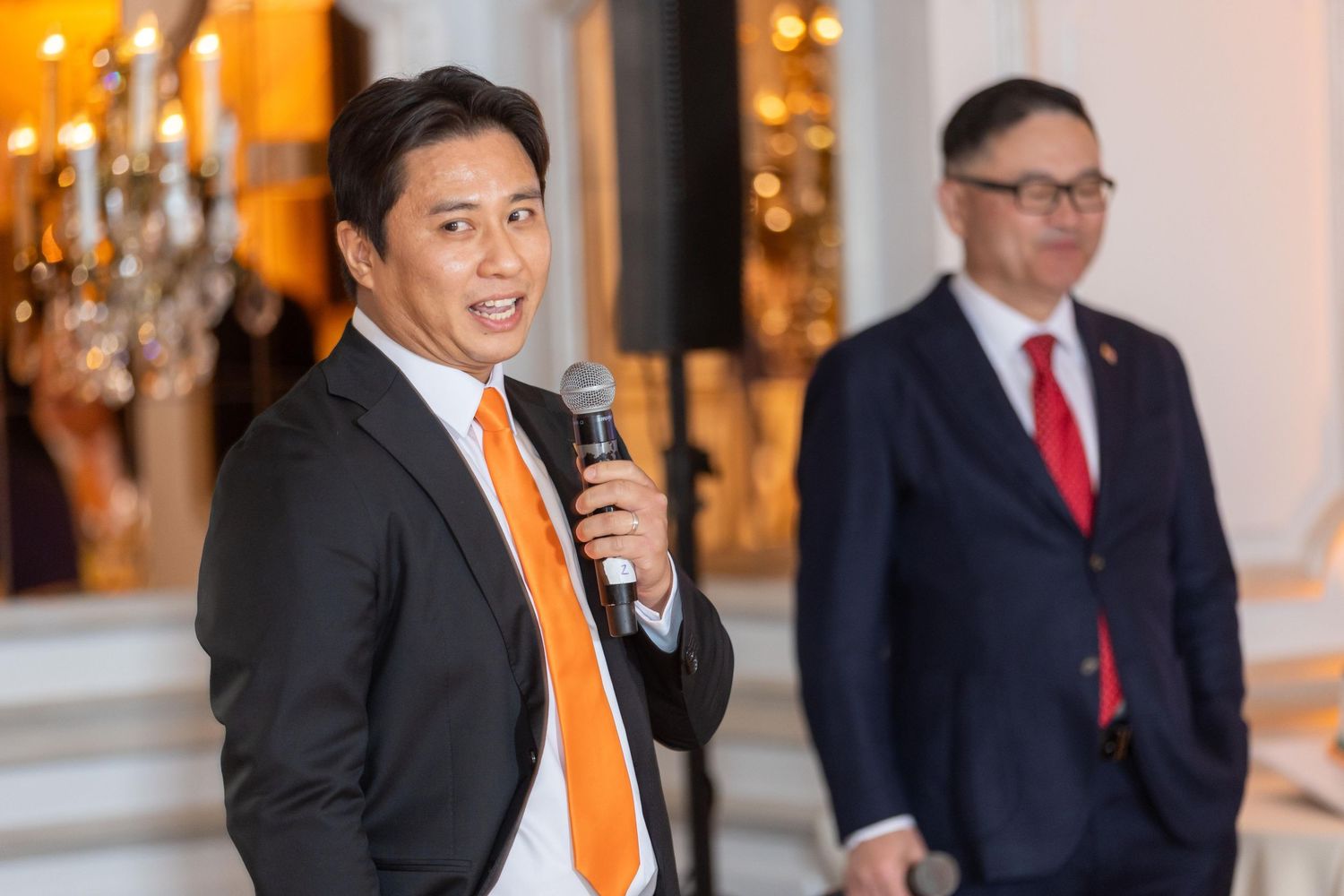 Man in black suit with orange tie speaks into microphone at formal event with colleague in background.