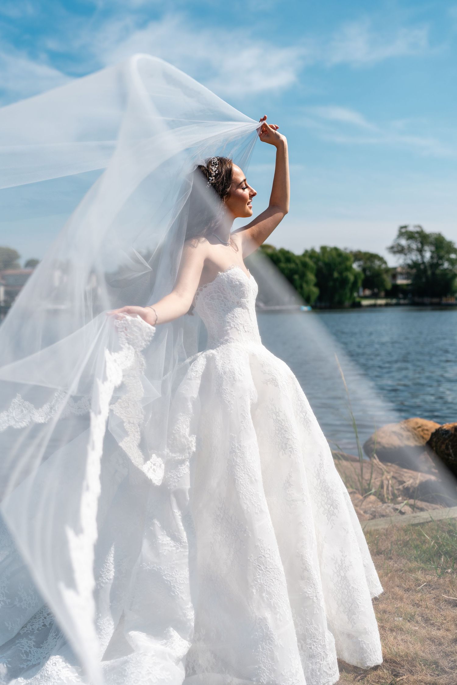 Bride in flowing white dress twirls gracefully by a lakeside on a sunny day.