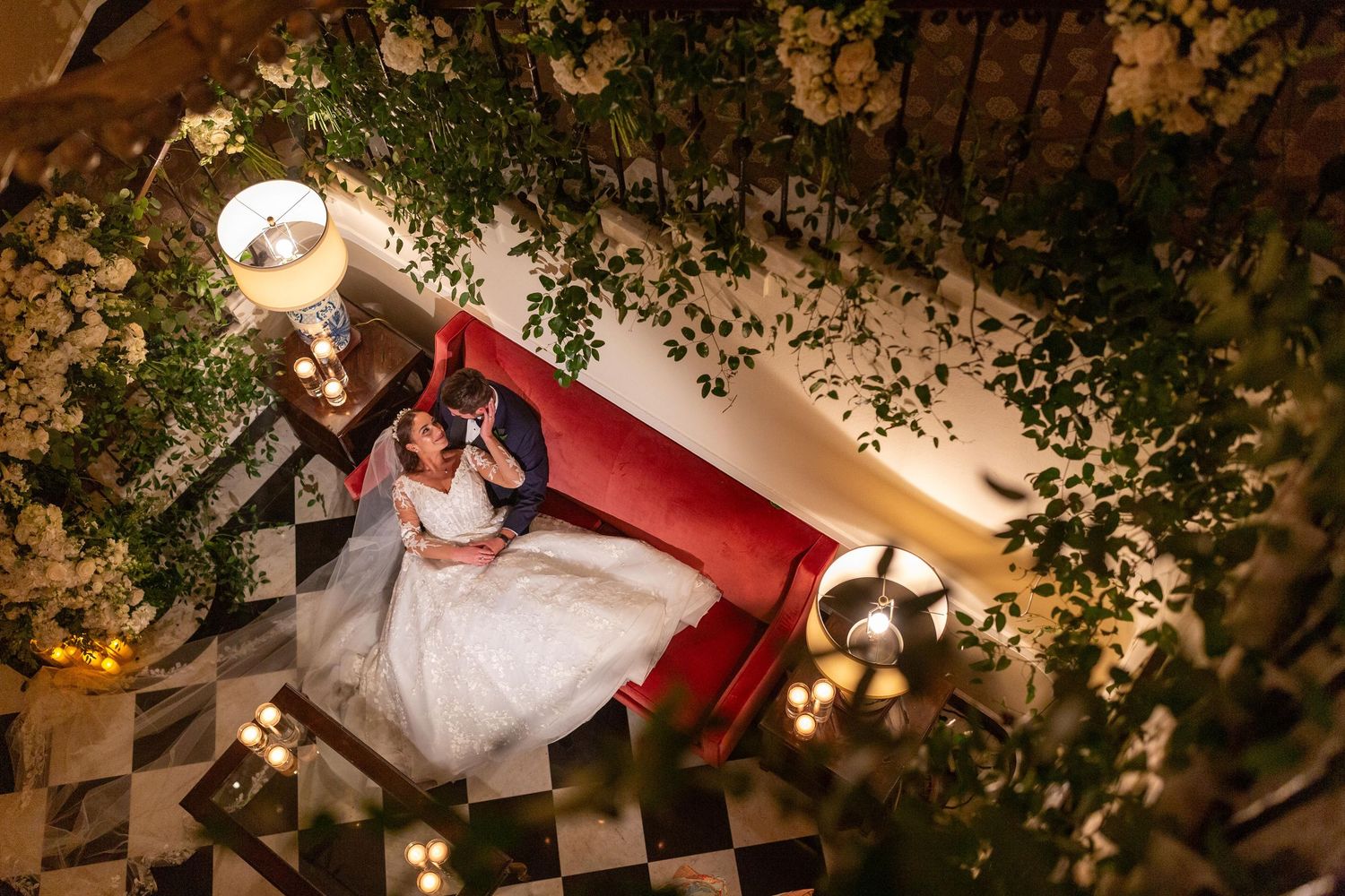 Wedding couple poses on red vintage furniture under twinkling garden lights.