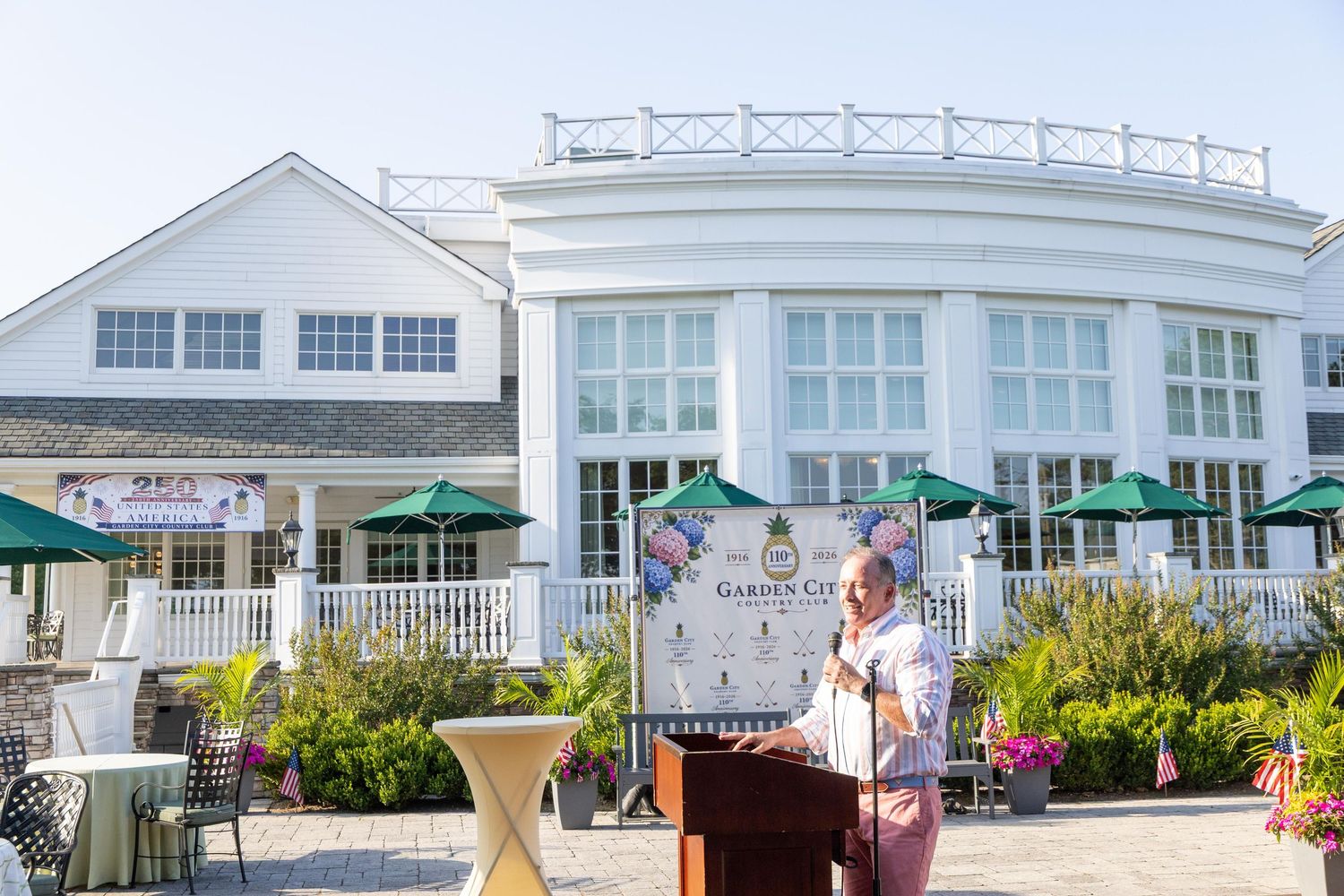 Speaker at podium outdoors near elegant white clubhouse with green umbrellas at a golf club event.