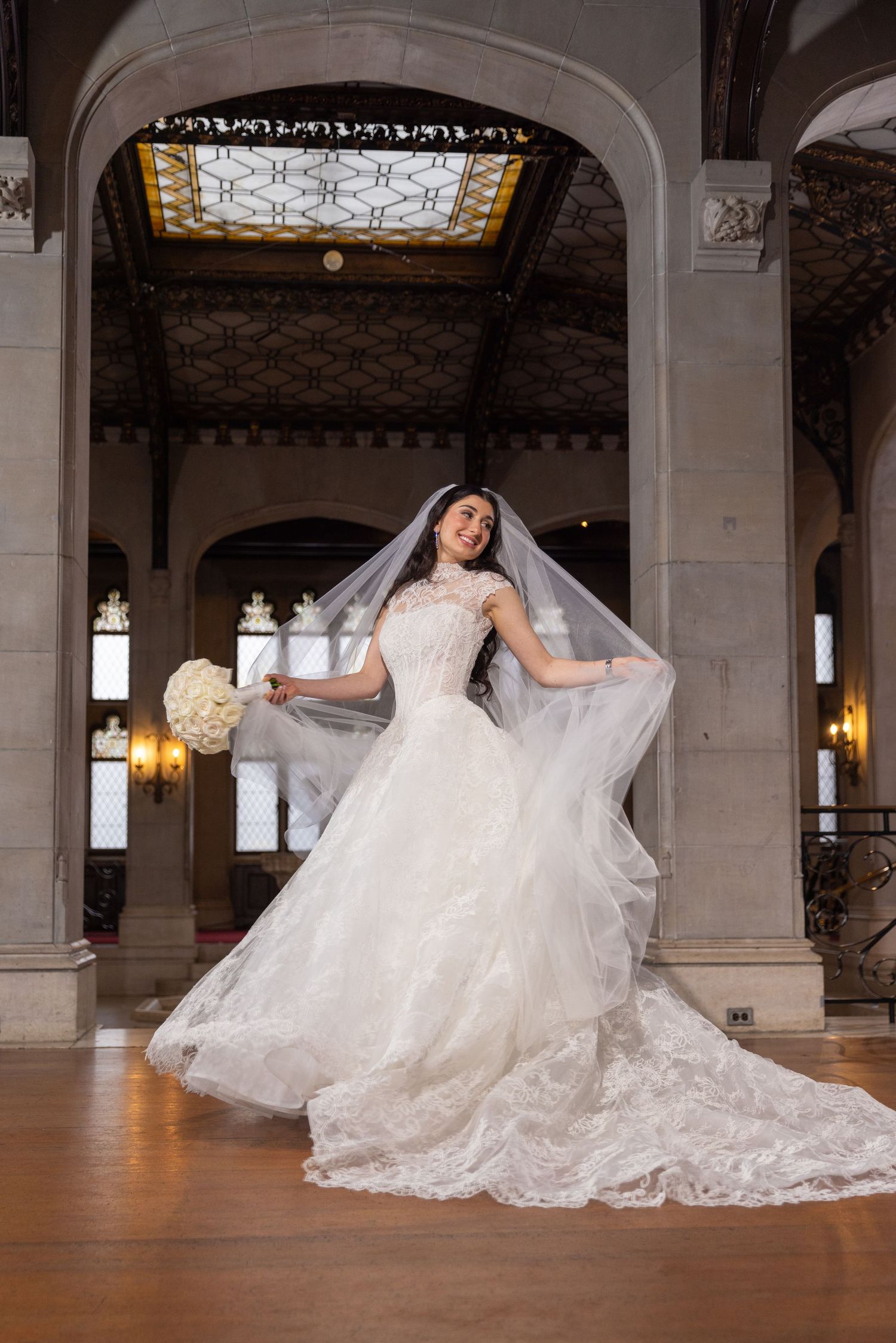 Bride in a flowing white lace wedding gown and veil twirling in an elegant ornate hall with stained glass ceiling.