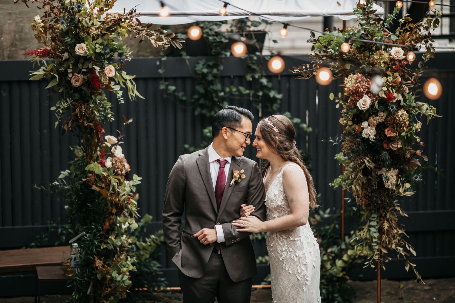 Wedding couple shares intimate moments under floral arch with string lights.