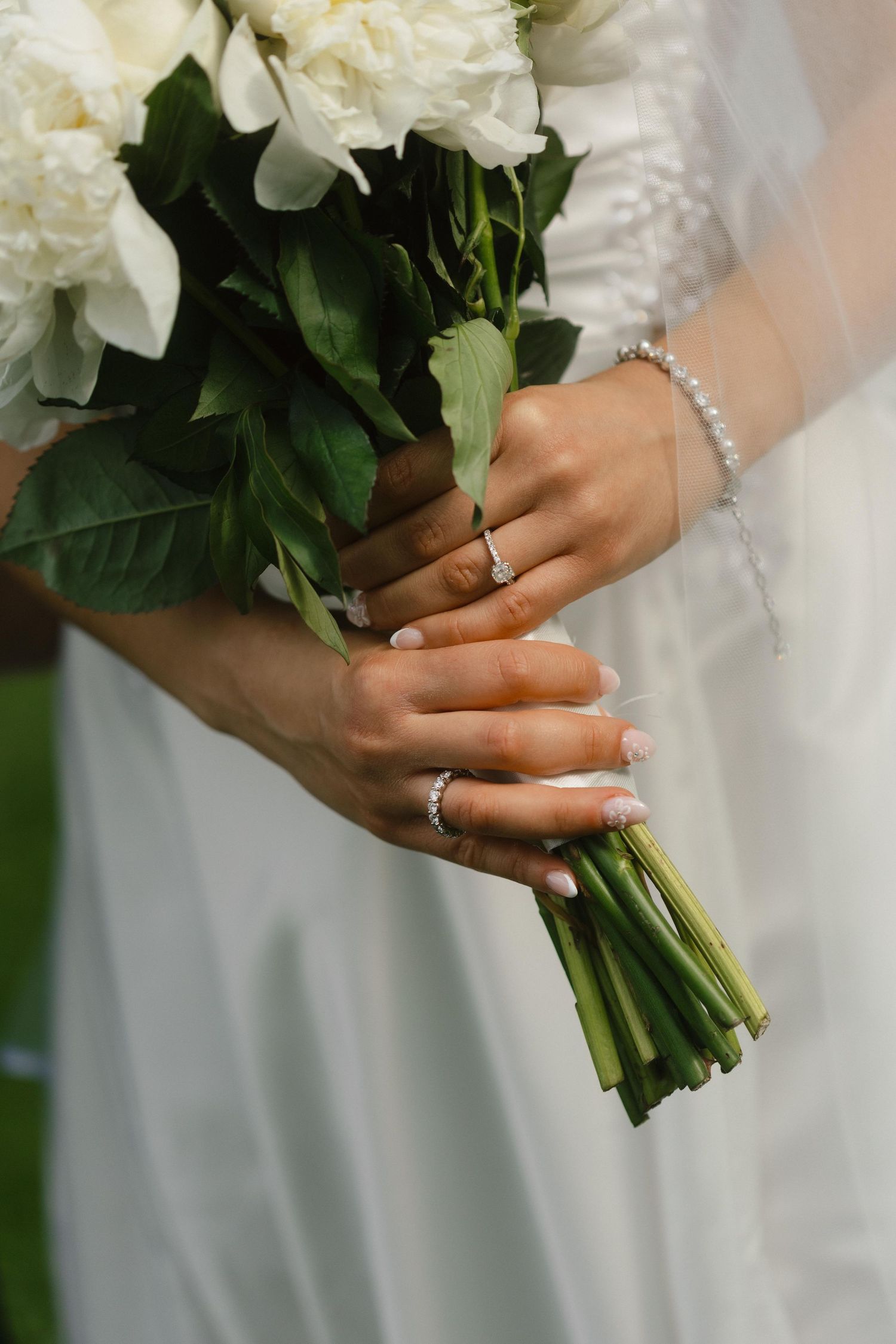 Close-up of white peony bouquets held by hands with wedding rings and bracelets.