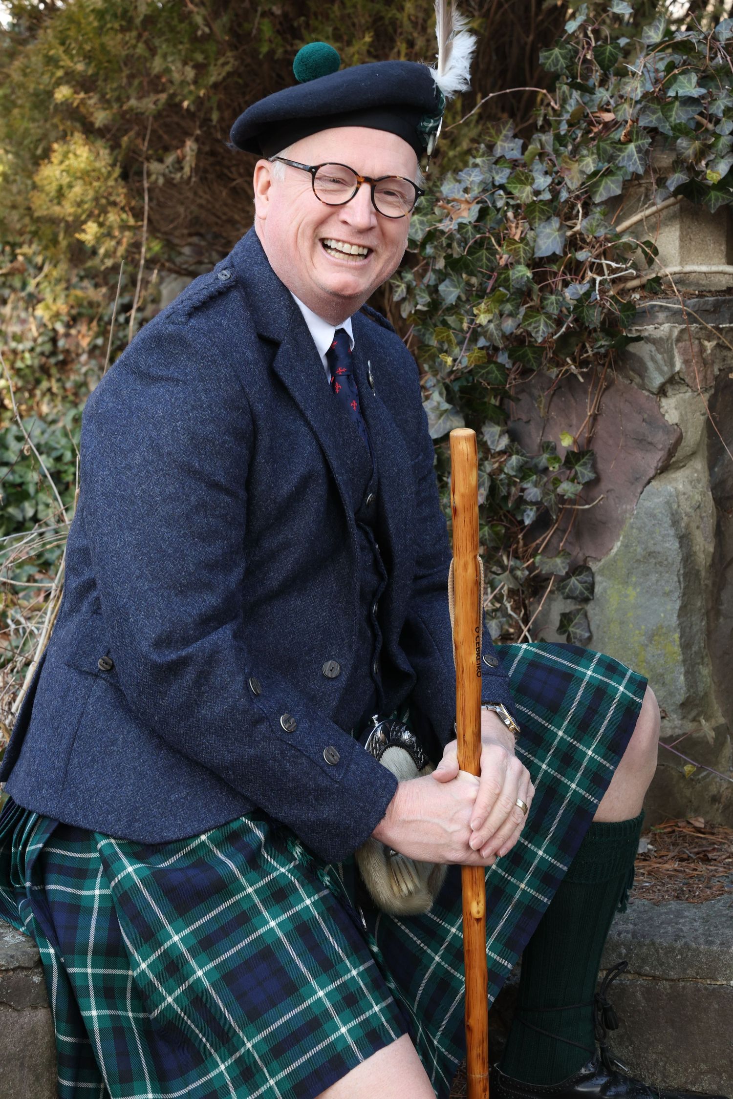 A person in traditional Scottish attire including a navy jacket, green tartan kilt, beret and glasses poses with a wooden staff.