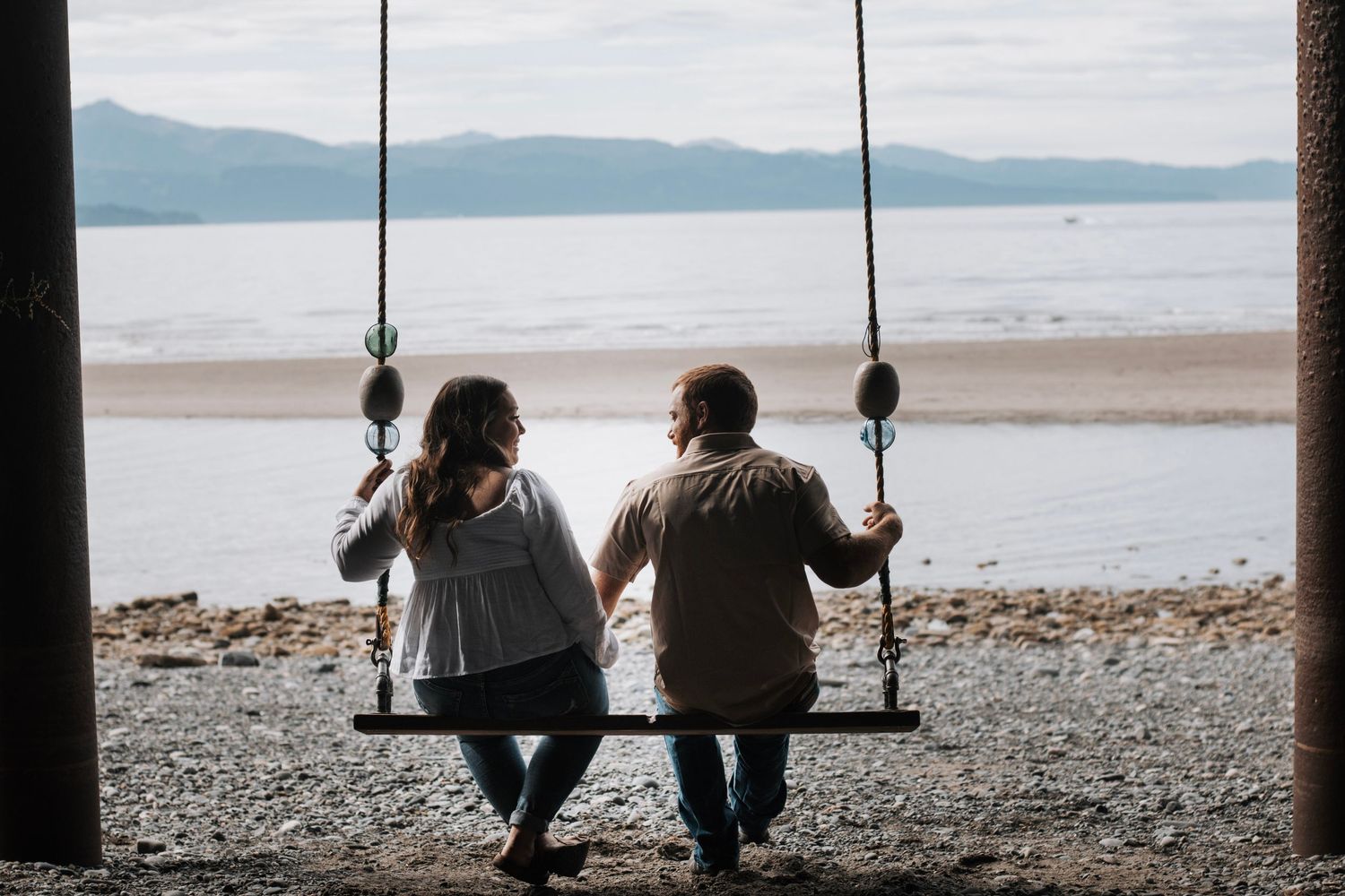 Silhouetted figures sit together on swings at sunset by a peaceful beach with distant mountains in Alaska