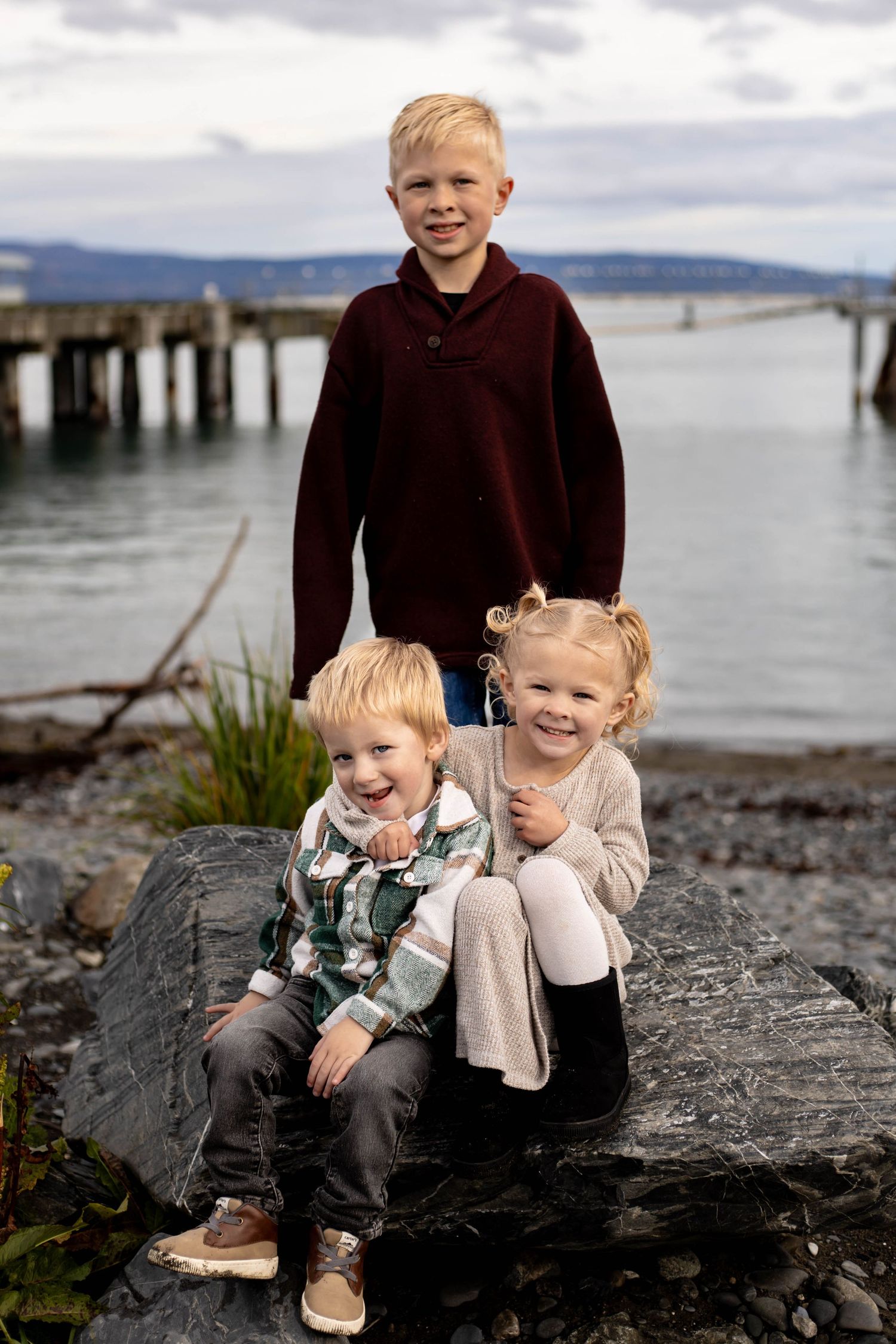 Children sitting together on rocky beach shore with cloudy skies and water in background.