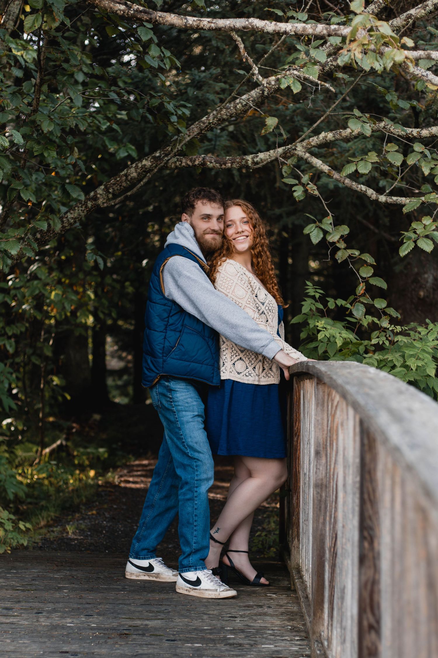 A couple shares affectionate moments while sitting on a wooden railing in a natural setting.