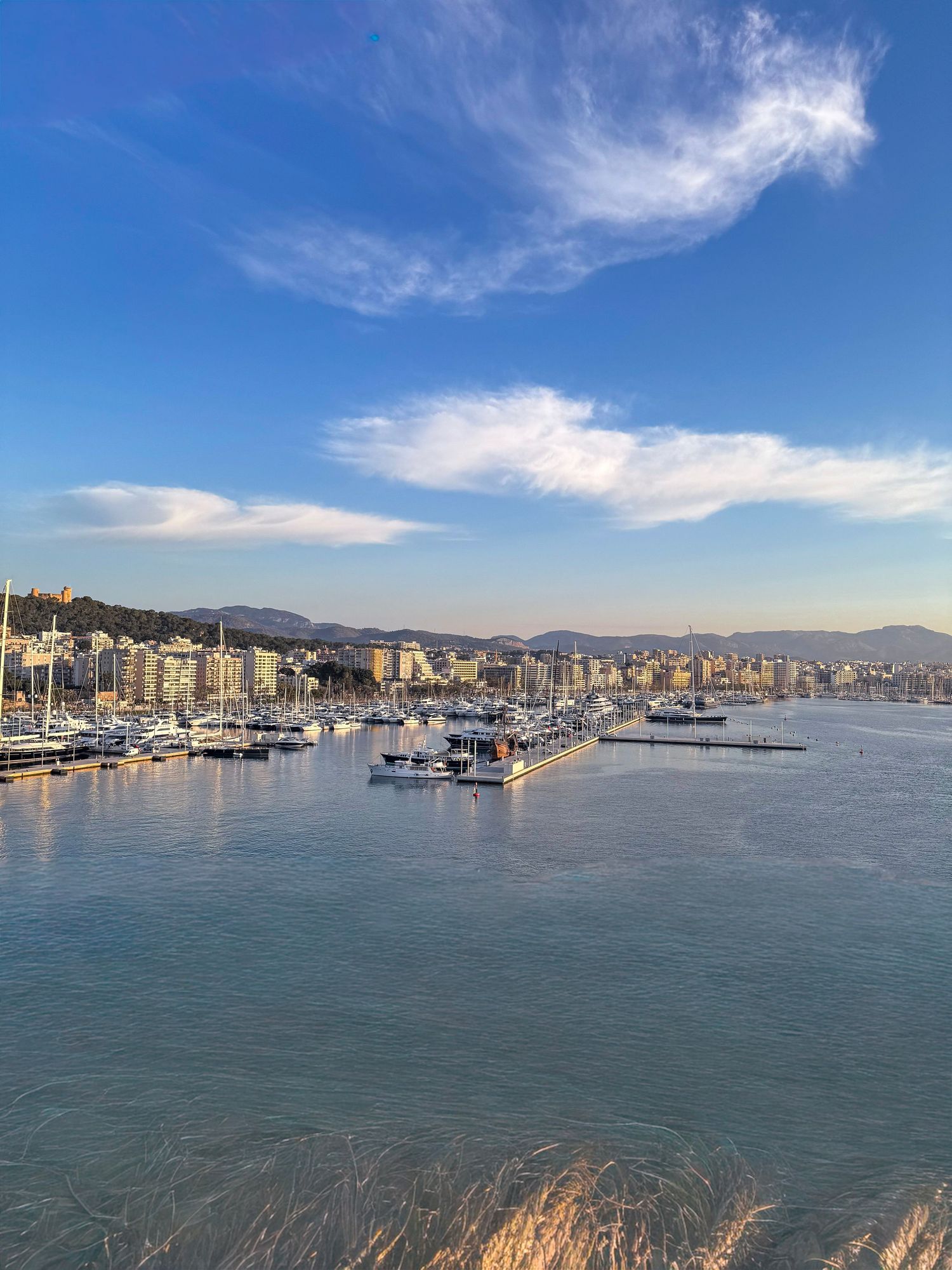 Panoramic view of a coastal cityscape with mountains, harbor, and boats against a blue sky with wispy clouds.