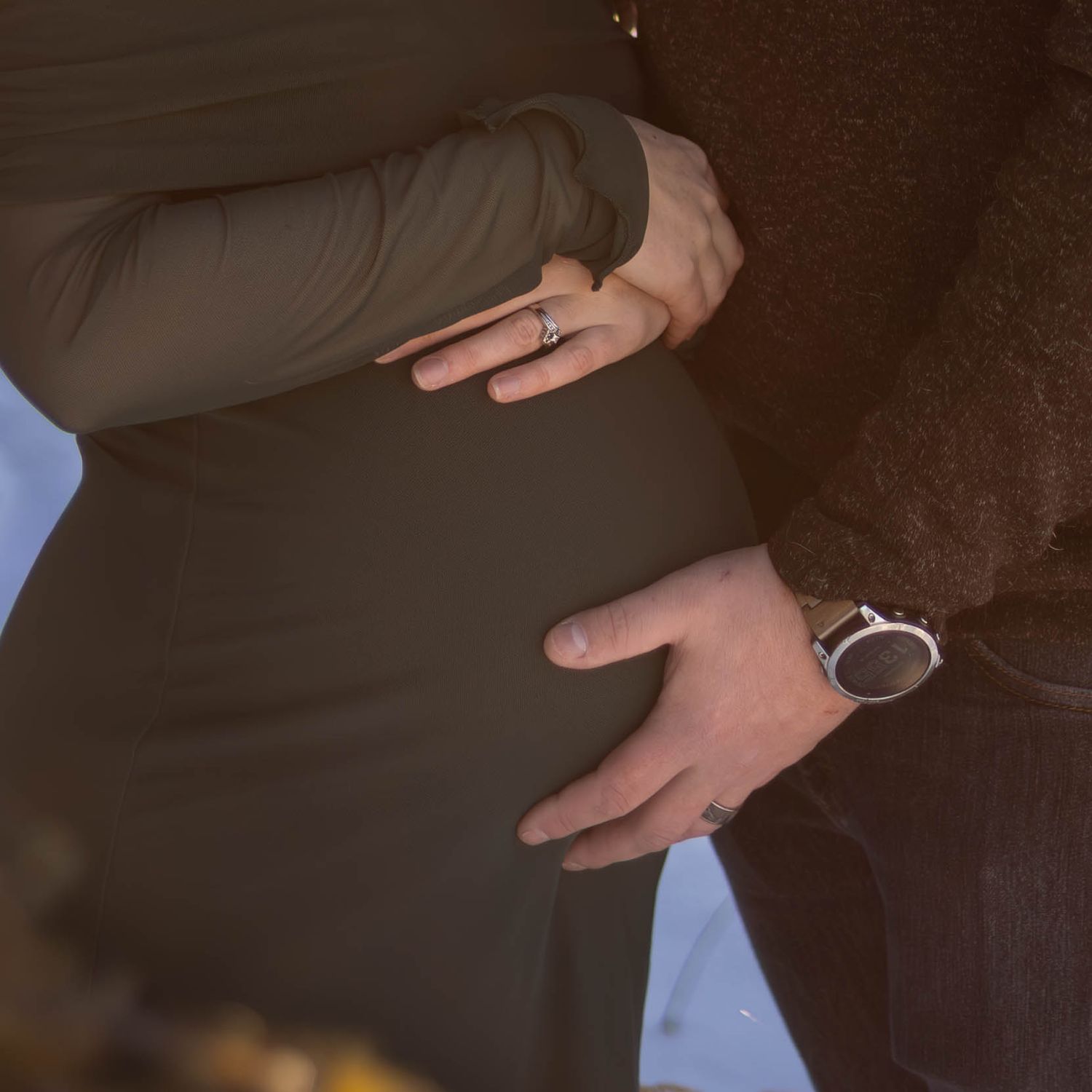 Couple cradling pregnant belly outdoors, woman wearing ring, man with watch, maternity photo shoot.