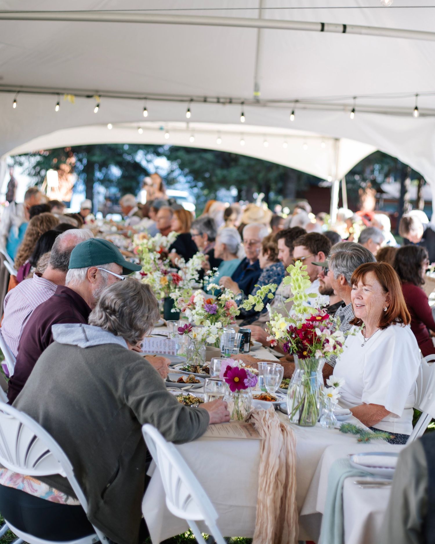 Guests dining under a white tent at an outdoor wedding reception with floral centerpieces and string lights.