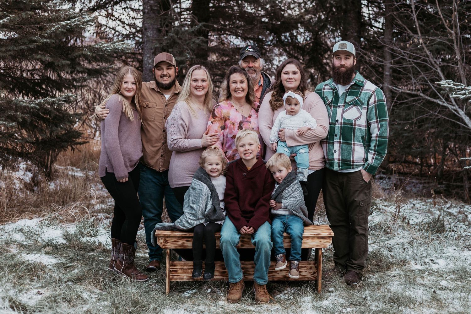 A family group sits together on a wooden bench in a snowy winter forest setting.