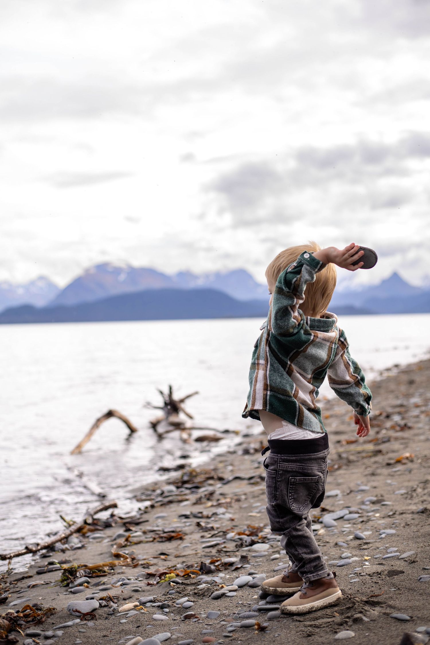 Child in winter clothing explores rocky beach with mountains and cloudy skies in the background.