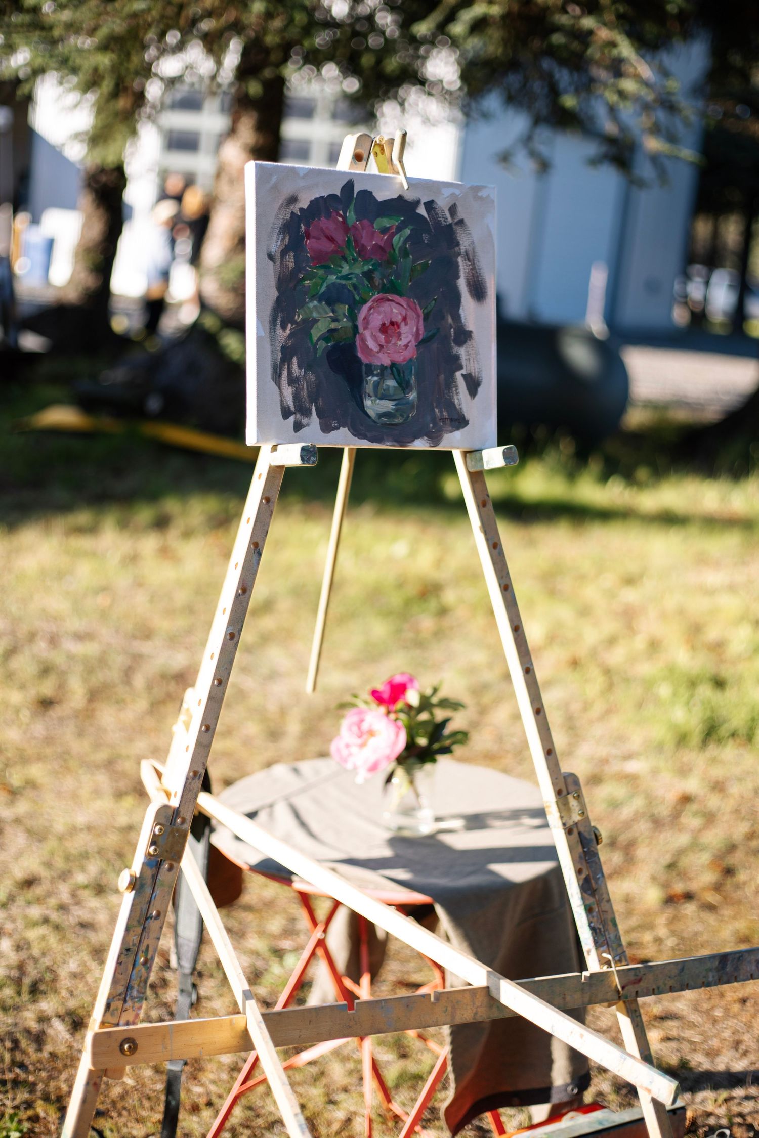 A floral painting on an easel outdoors with a pink flower bouquet on a table in the background.