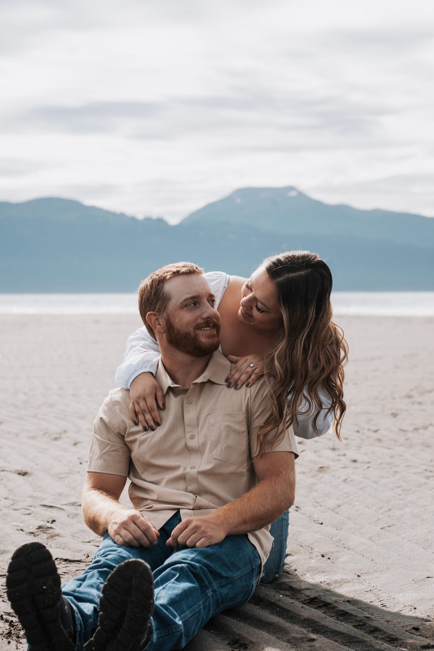 Couple sharing affectionate moment while sitting on sandy beach with mountains in background.