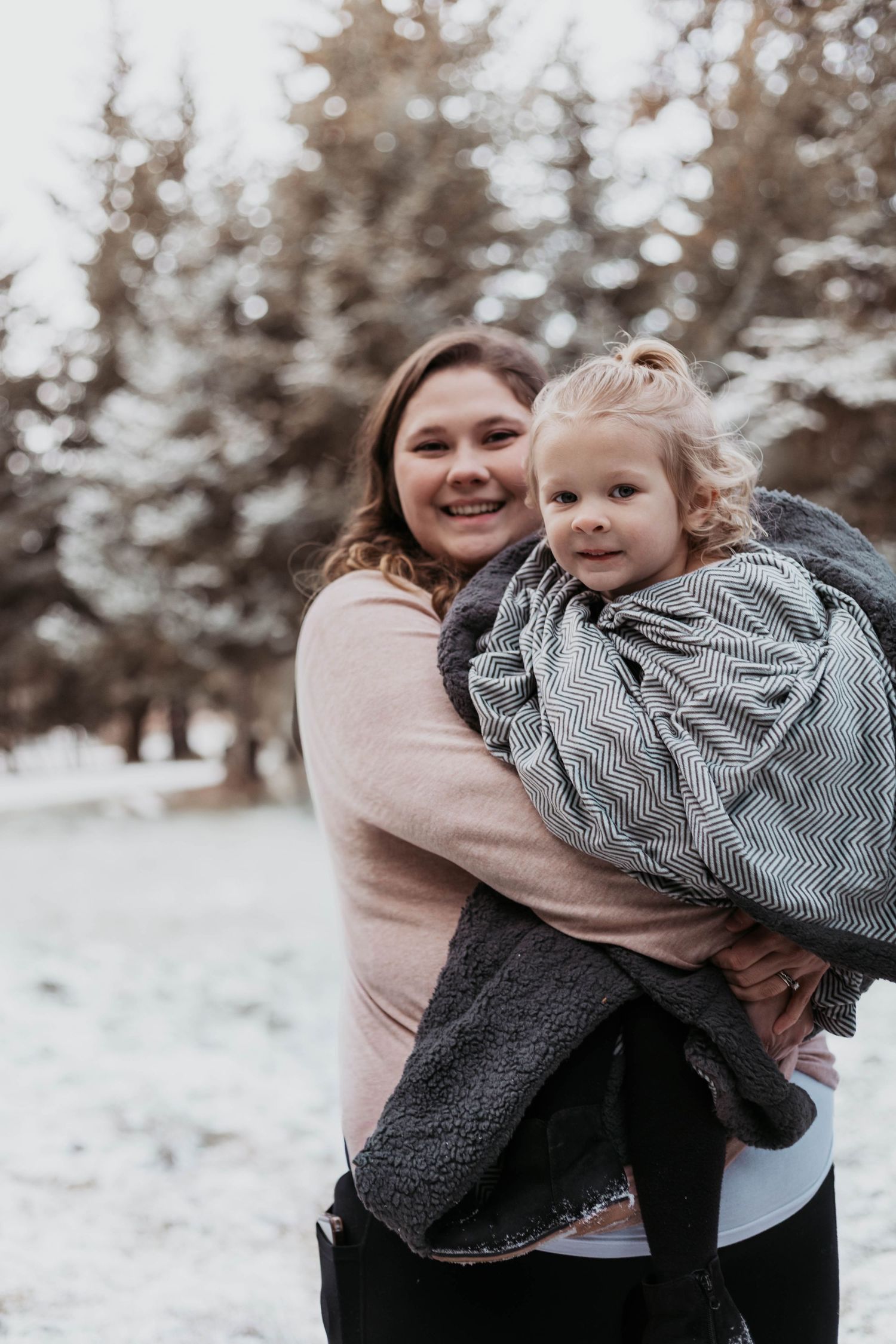 A smiling woman holds a young child wrapped in a blanket outdoors in a snowy winter forest setting.