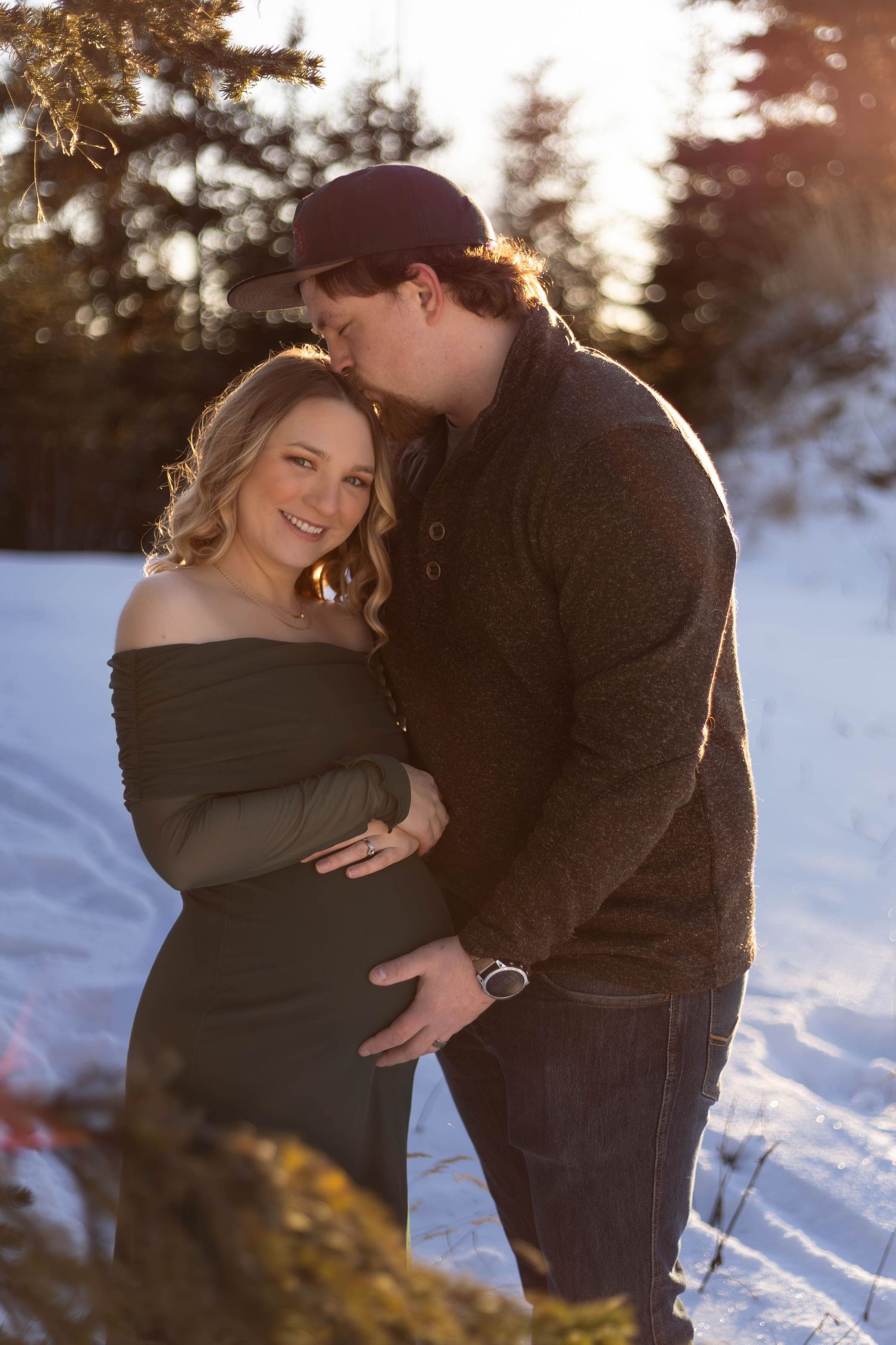 Pregnant couple embracing outdoors in snow, man kissing woman's head at golden hour, wearing winter clothing.