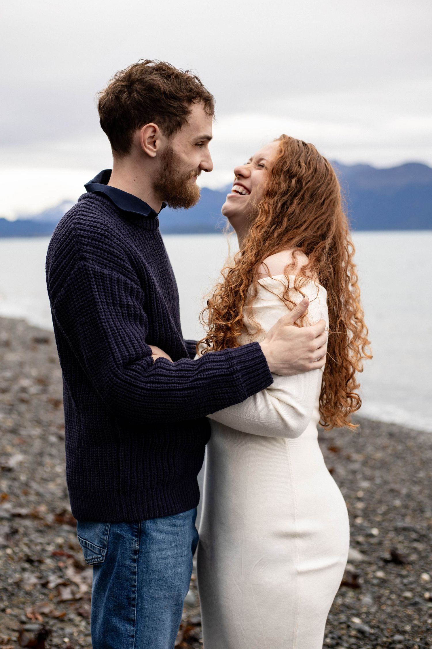 A couple embraces on a rocky beach shore with mountains and cloudy skies in the background.