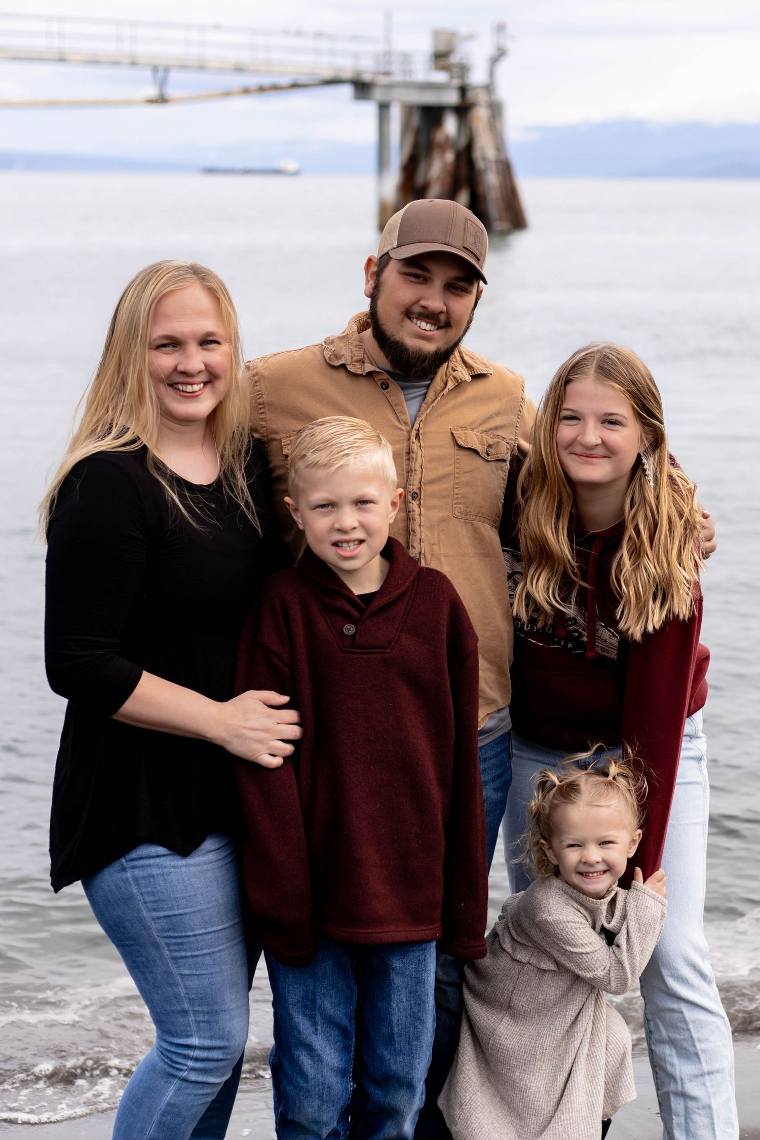Family of five smiling together near a waterfront pier on a cloudy day, wearing casual fall clothing.