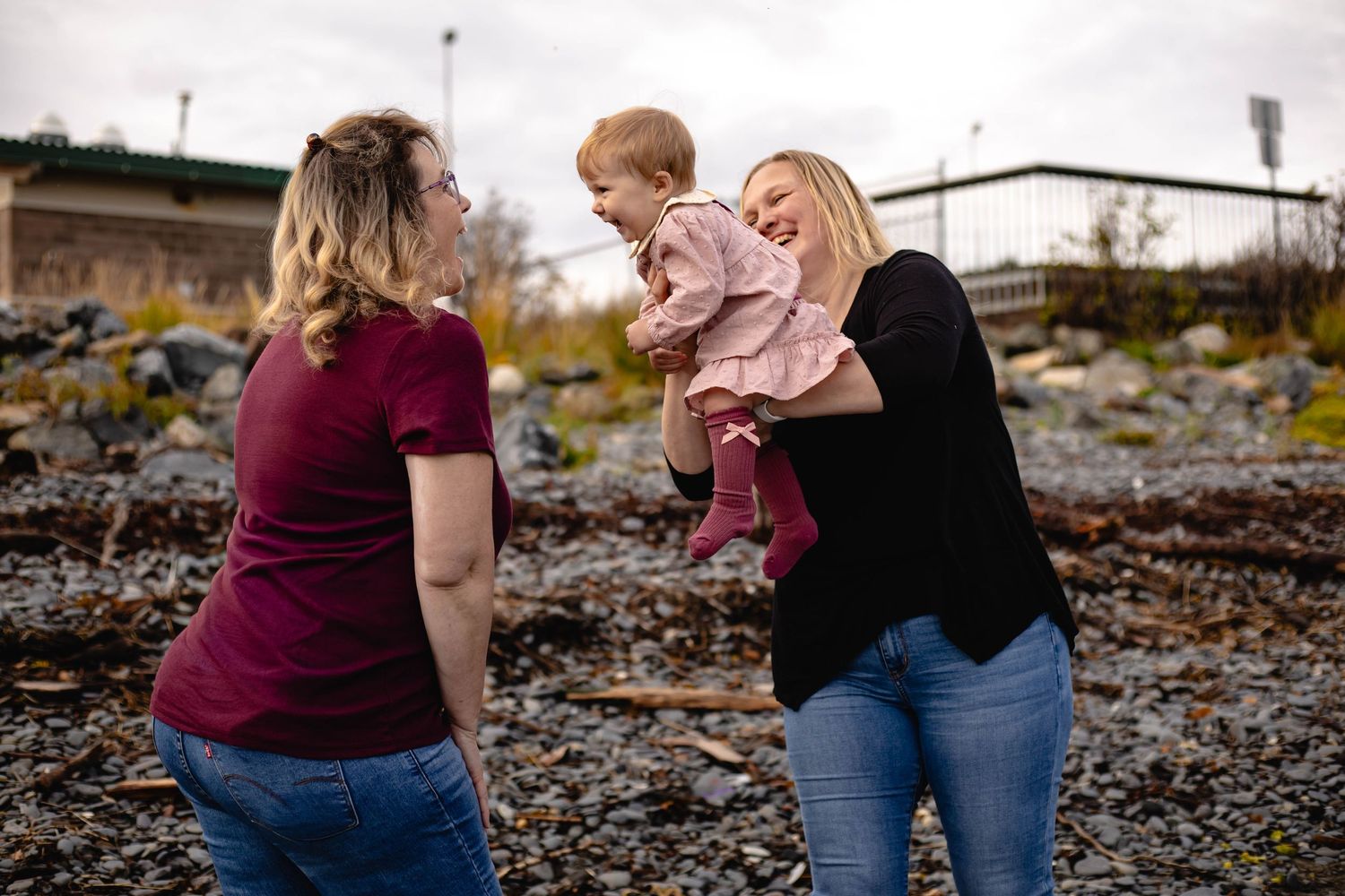 Two women smiling and interacting with a baby girl outdoors on a rocky beach setting.