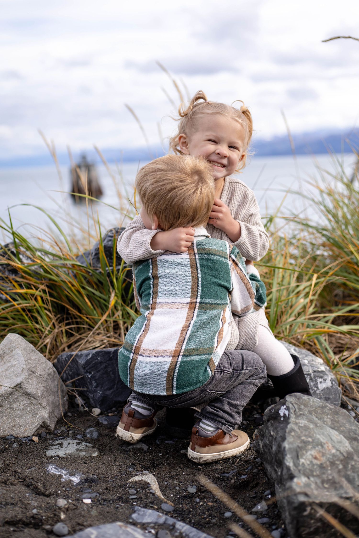Young child plays on coastal rocks while wearing a plaid vest on a cloudy day.