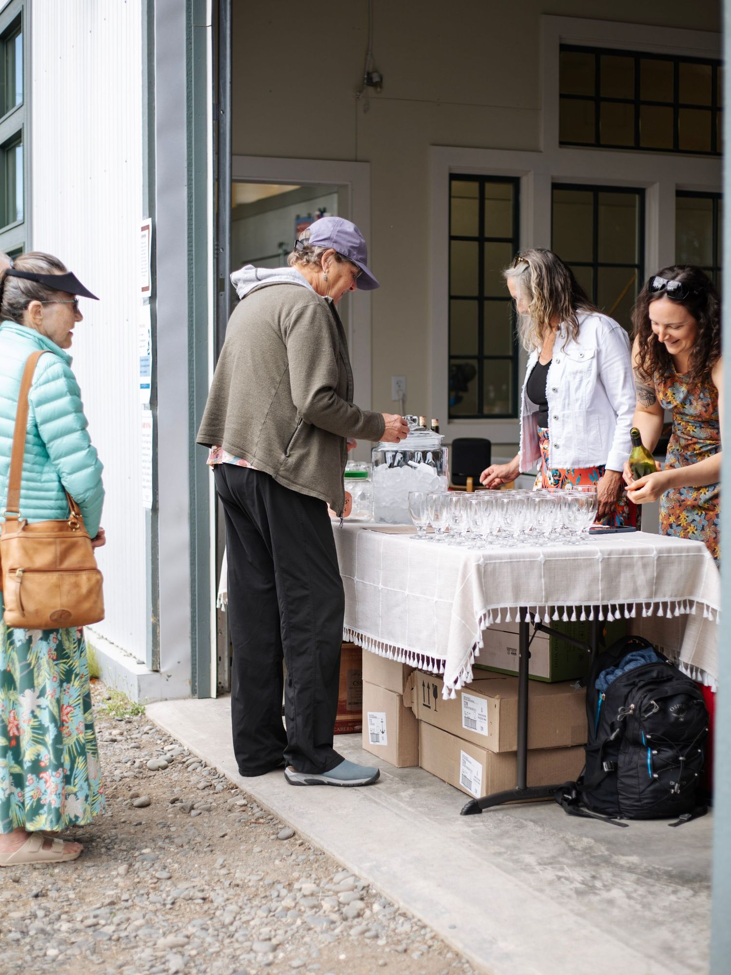 People gathered around an outdoor table with wine glasses at a winery event near a white building.
