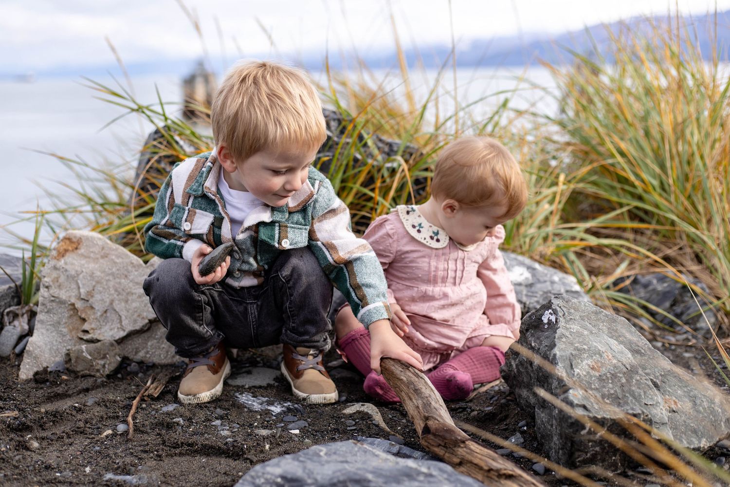 A group of children explore and play together in tall grass and sand near the beach on a cloudy day.