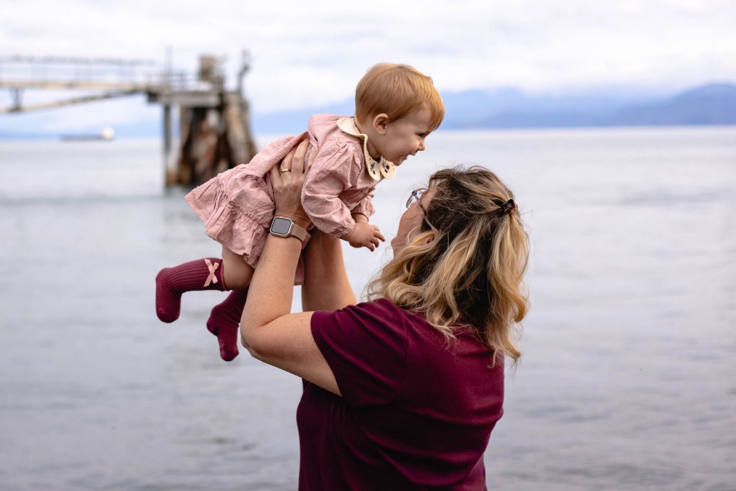 A parent lifts a child up playfully at the beach with mountains and water in the background.