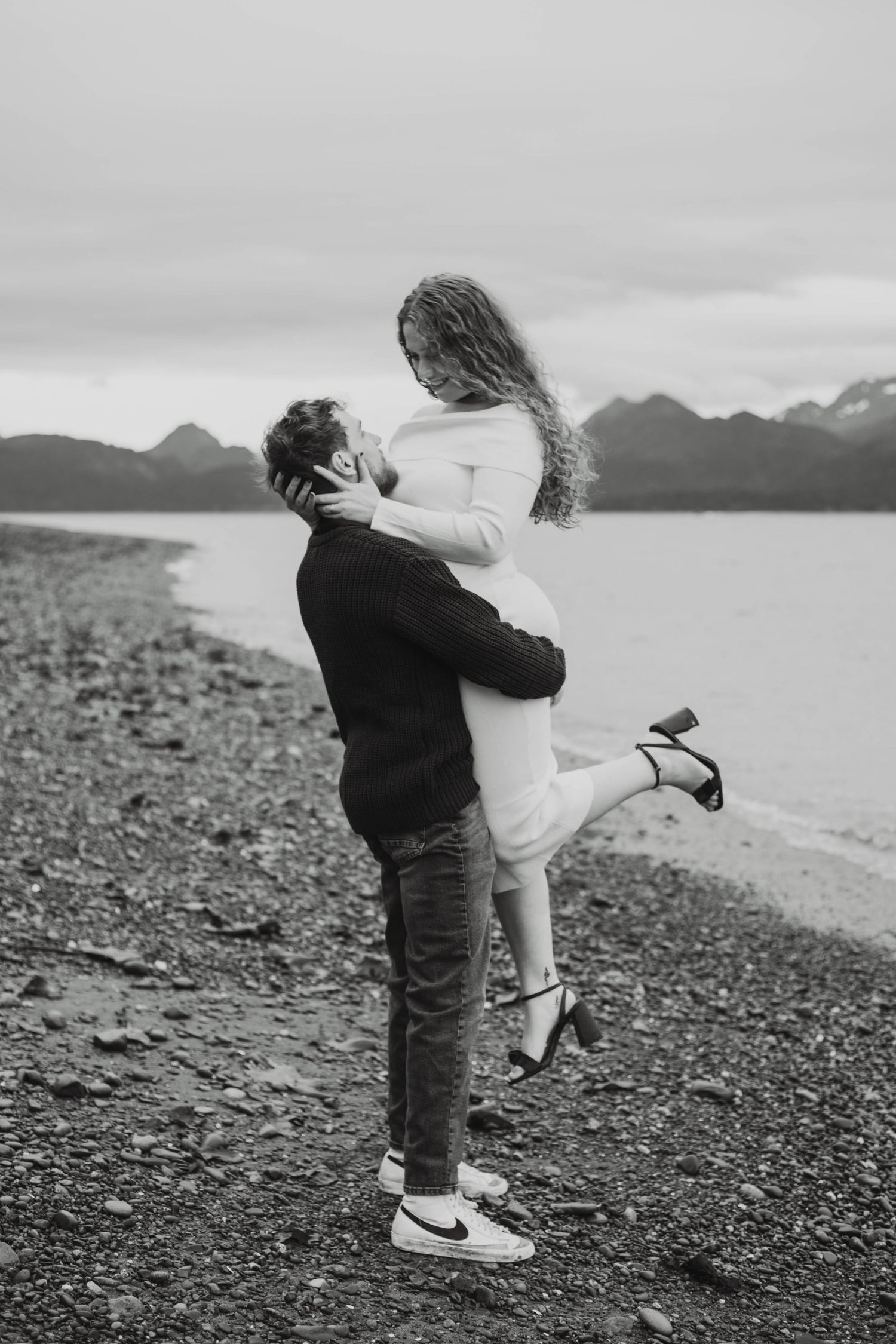 A black and white photo of a couple sharing an intimate moment on a beach with mountains in the distance.