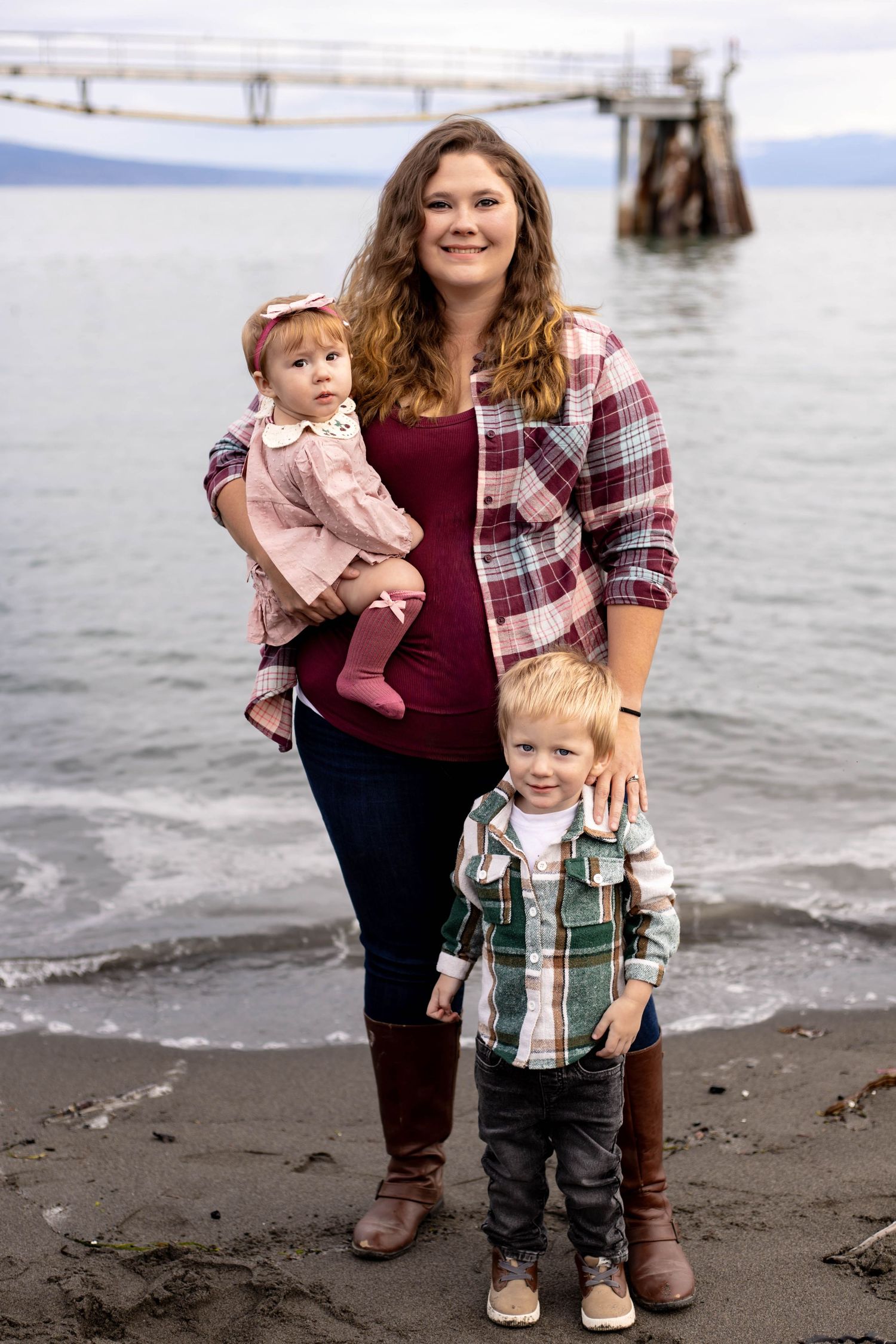 A young mother poses on a beach with her toddler son and baby daughter, with a pier and ocean in the background.