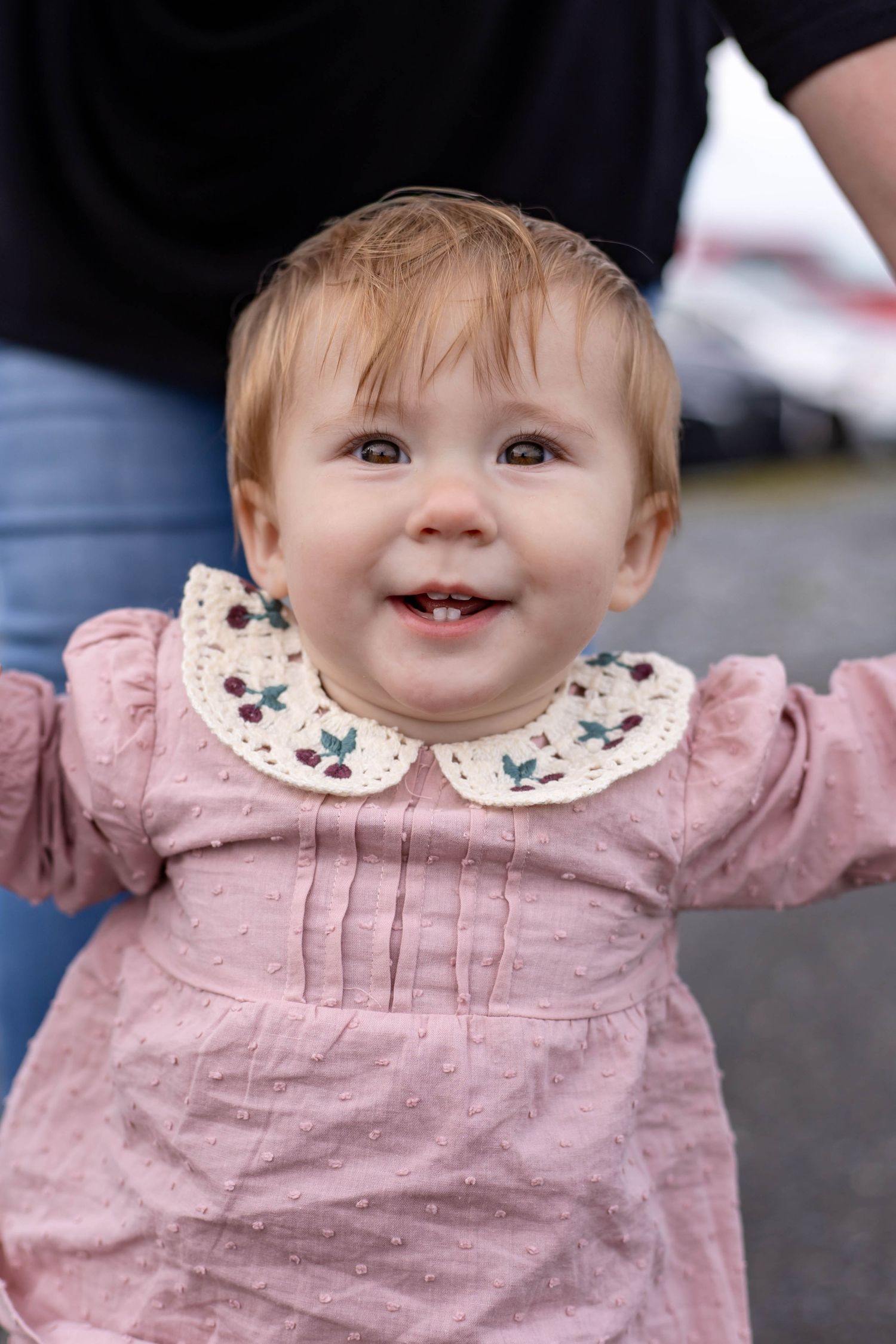 A small child in a pink dress with floral collar smiles brightly.