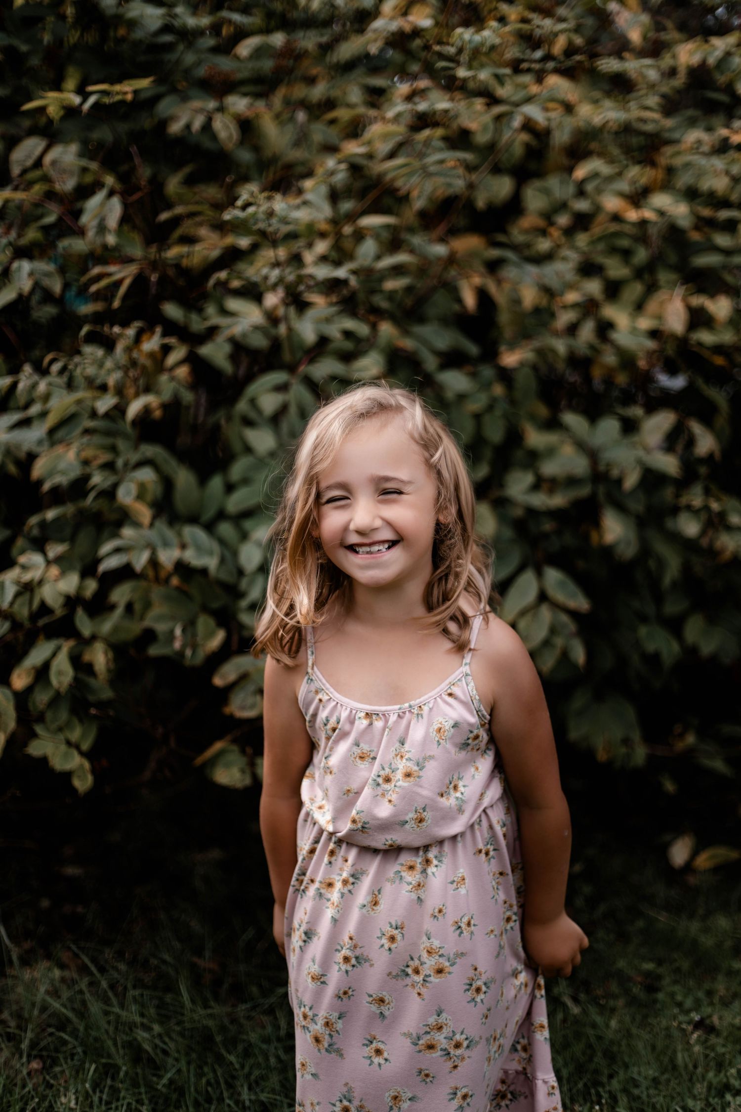 A young child wearing a floral summer dress stands against a backdrop of lush green foliage.