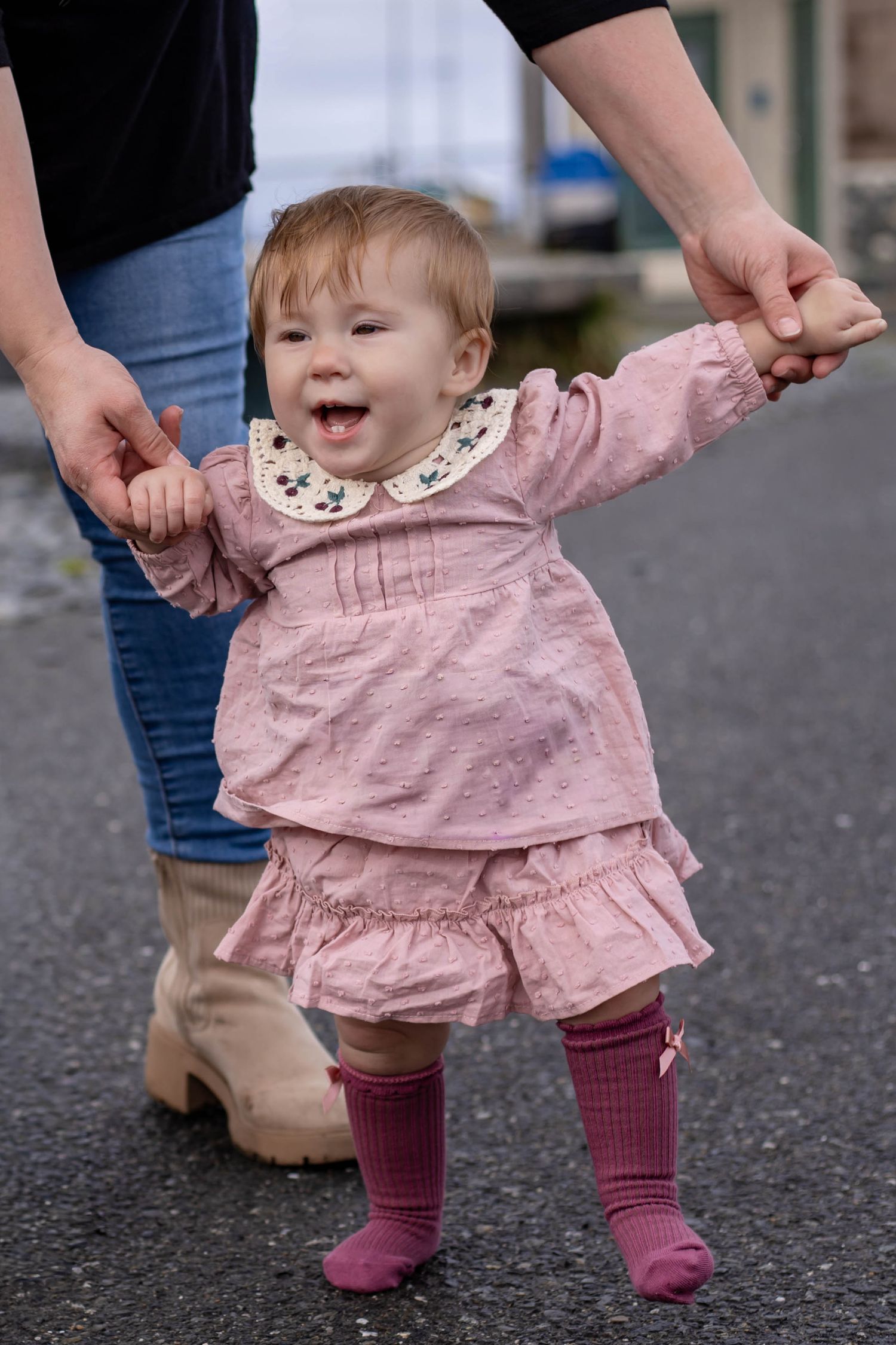 Toddler girl in pink dress and socks takes first steps outdoors, held by adult hands for support.