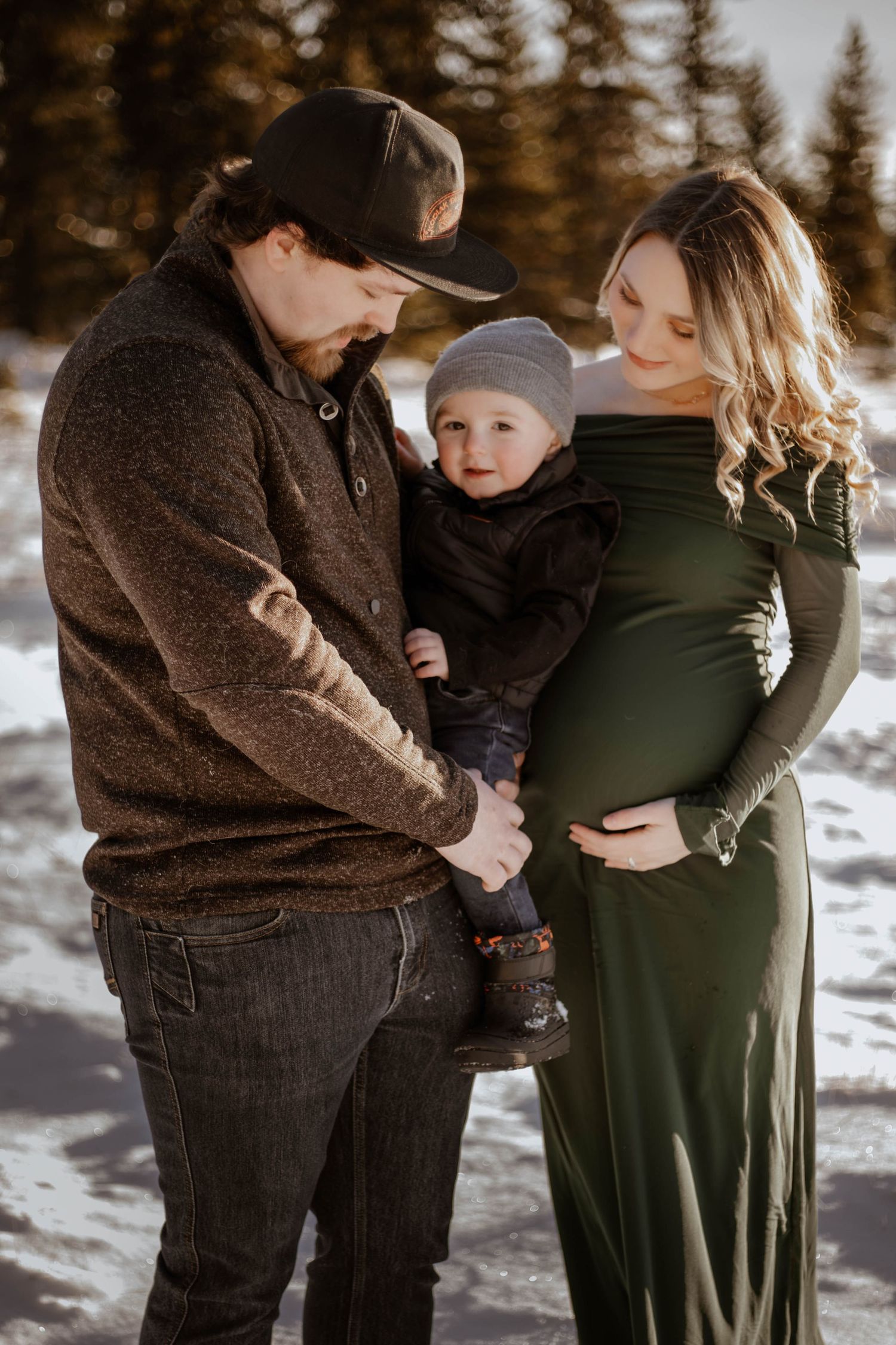 Pregnant woman in green maternity gown with husband and toddler son pose for winter family maternity photos in snow.