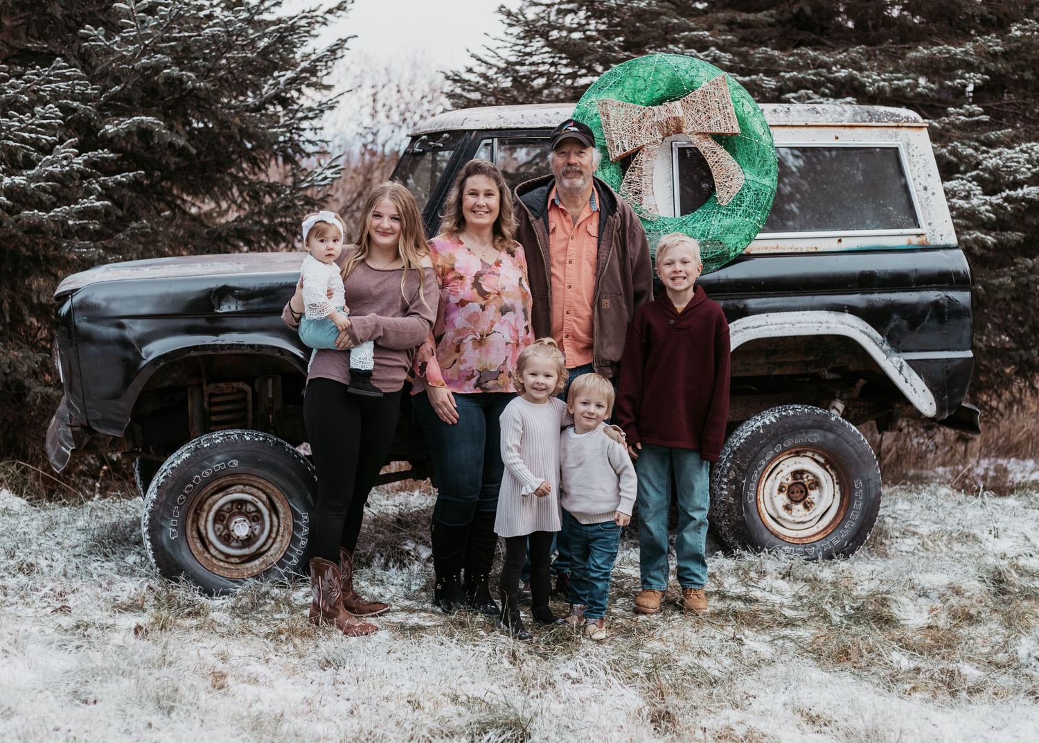 Family of six posing in front of a vintage Ford Bronco decorated with a Christmas wreath in a snowy field.