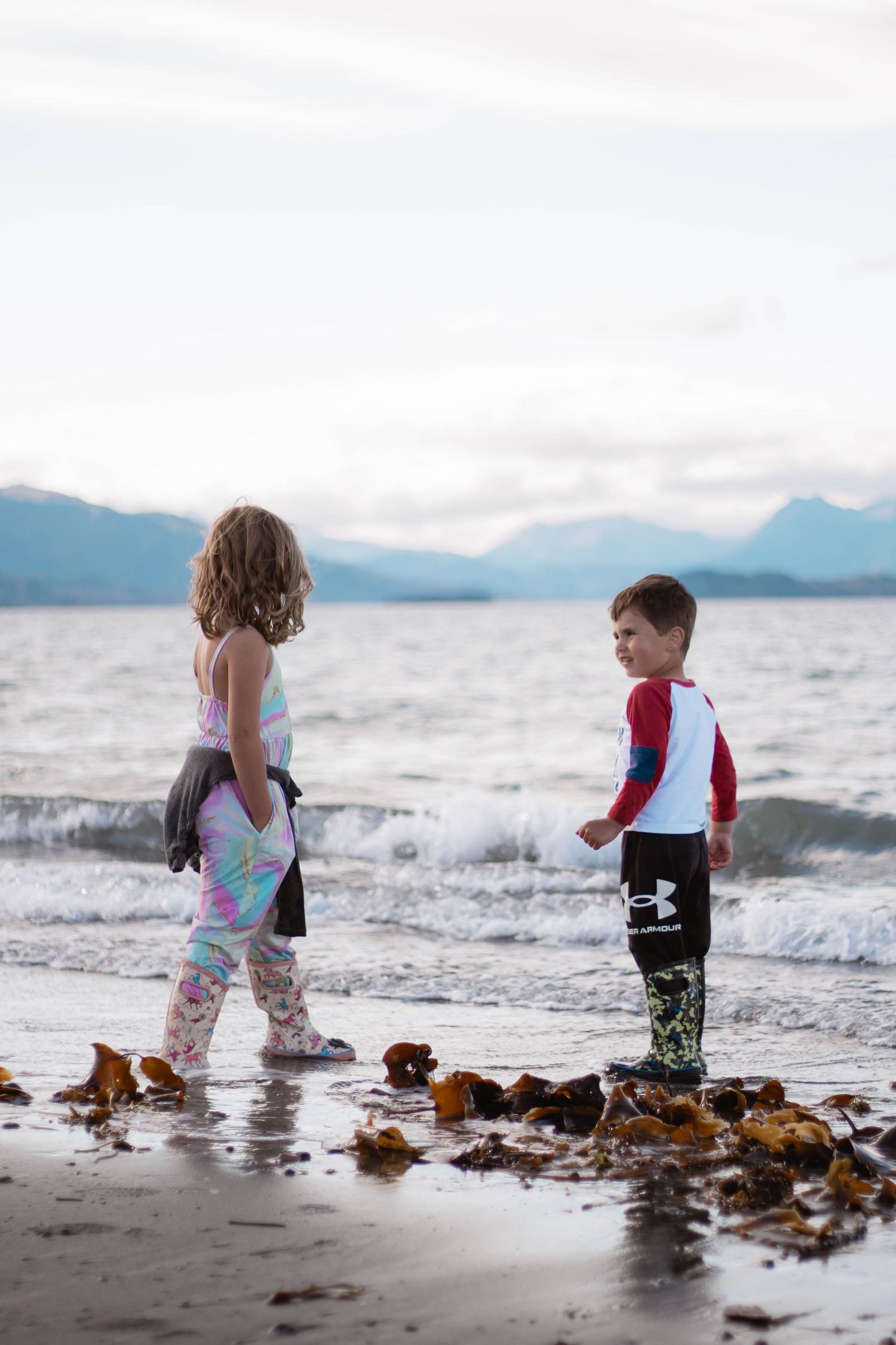 Two children play together at the beach's edge among seaweed and waves with mountains visible in the background.