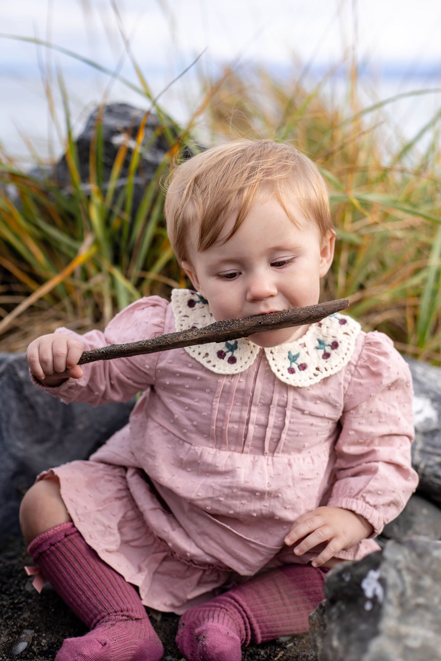 Baby in pink dress and burgundy tights plays with a stick while sitting on rocks outdoors.