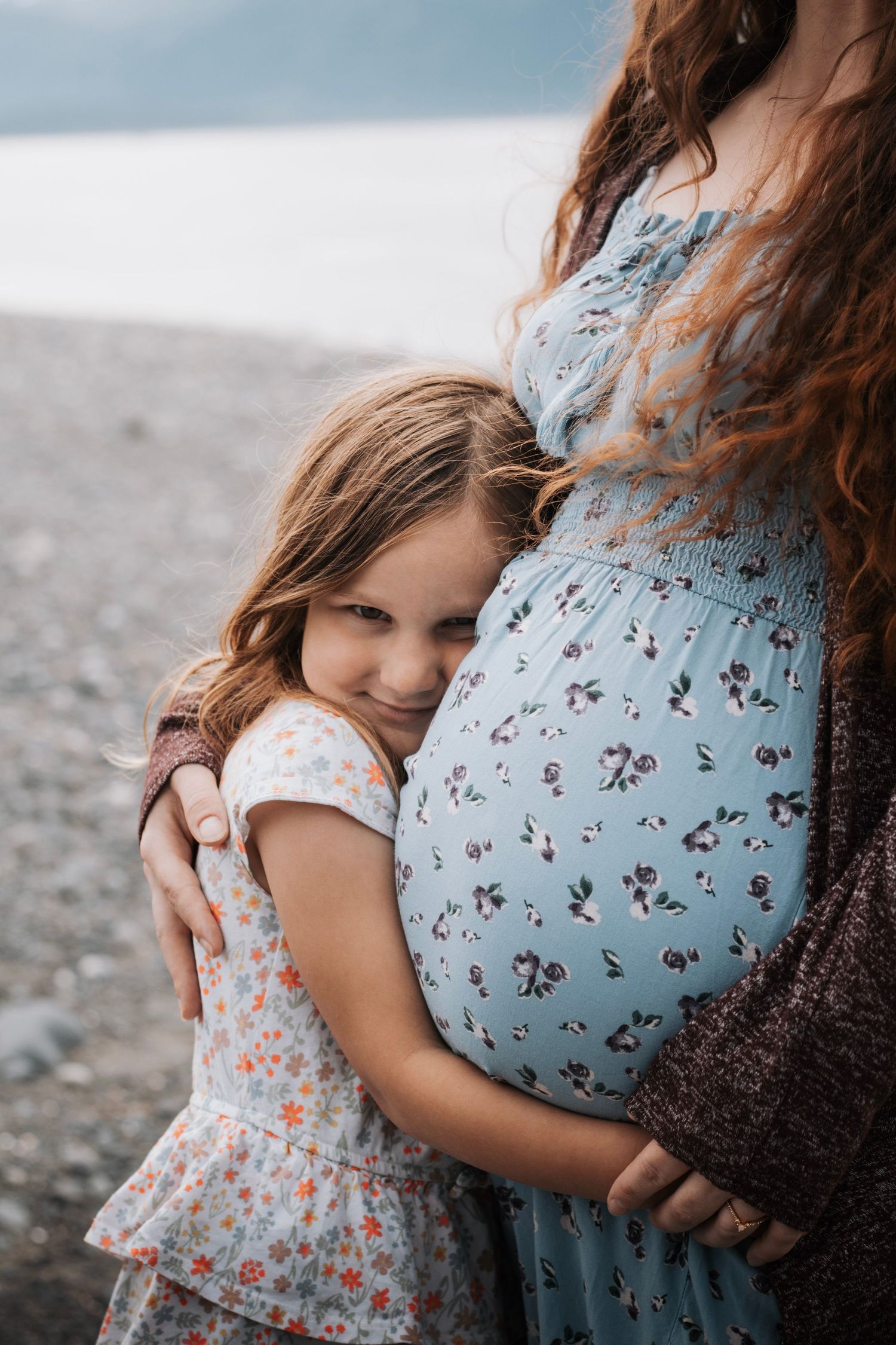 A young girl hugs her pregnant mother's belly on a rocky beach, both wearing floral dresses.