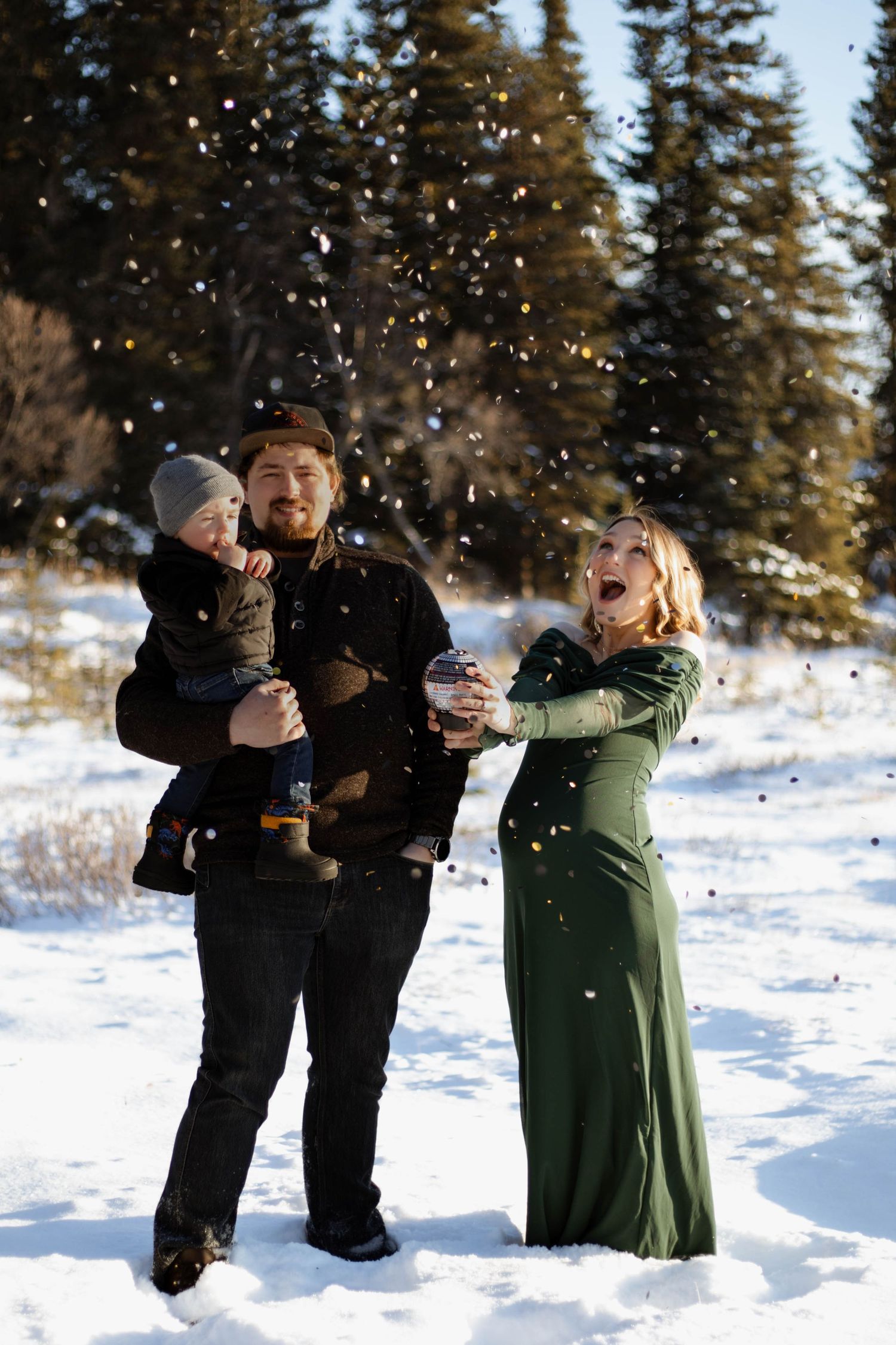 Family in snowy forest, man holding baby, pregnant woman in green dress, confetti flying, winter gender reveal.