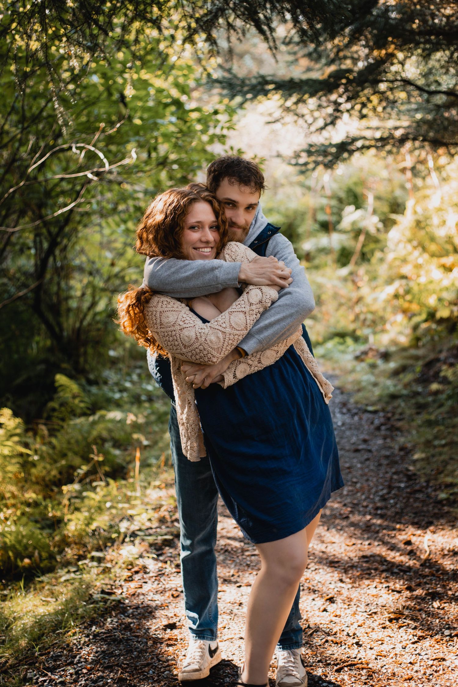 A couple hugs and dances together on a sunlit wooded path covered in fallen autumn leaves.