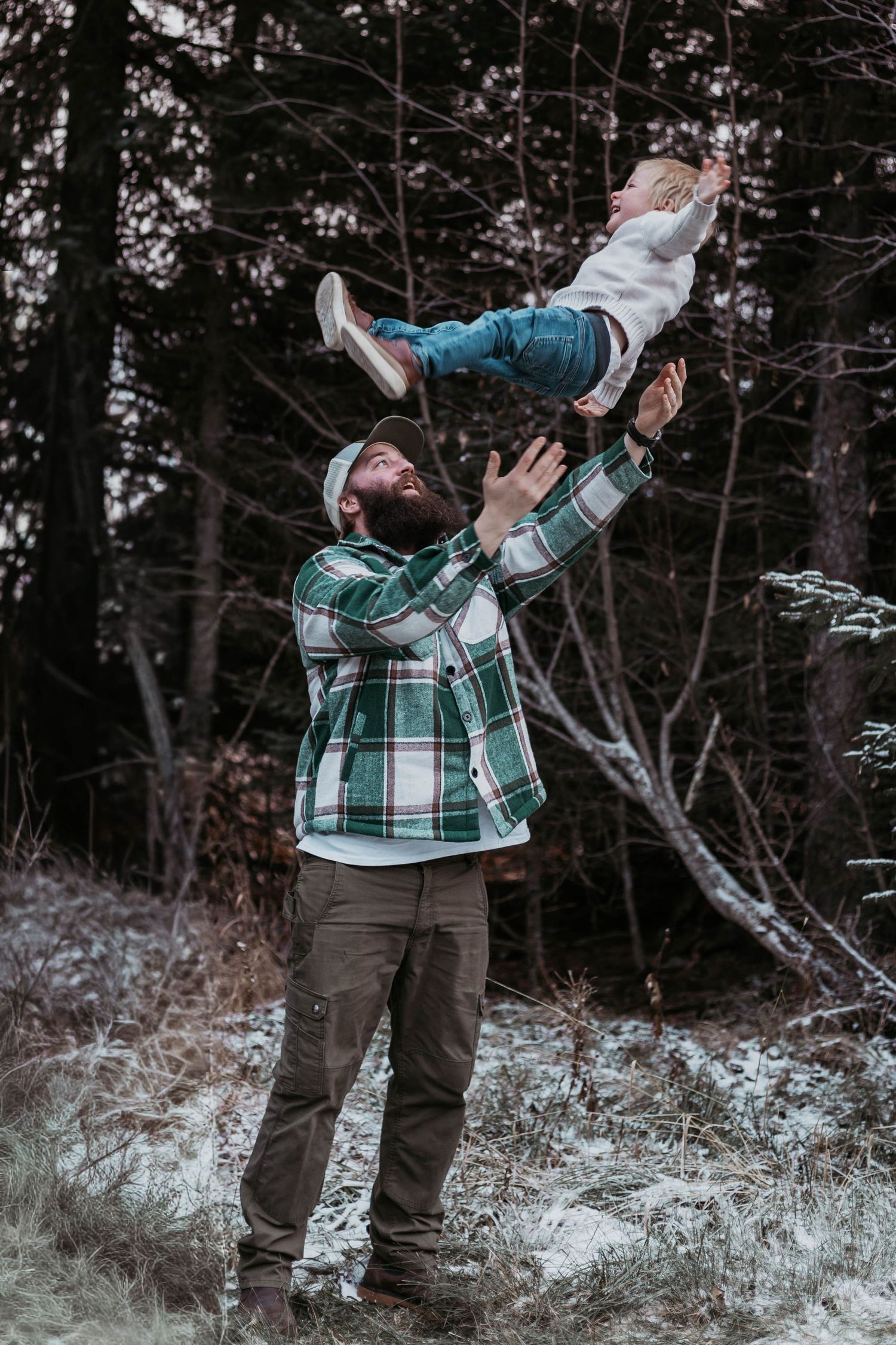 A bearded man in a plaid shirt joyfully tosses a young child into the air in a snowy winter forest.